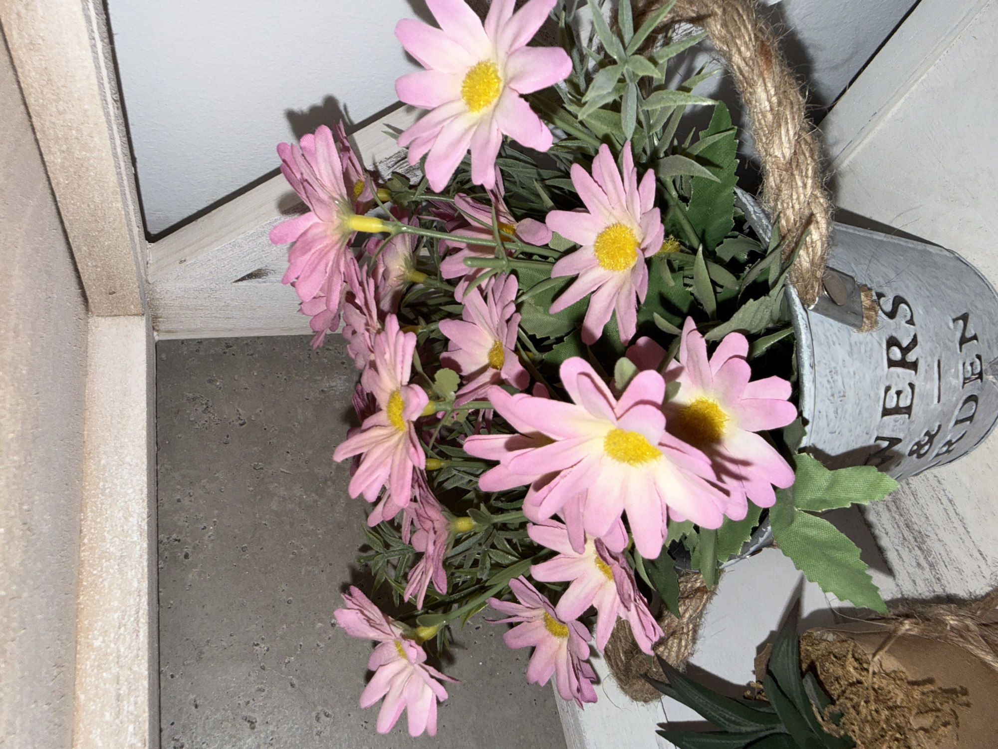 A close-up shot showcases a cluster of artificial pink daisies with yellow centers, artfully arranged in a weathered, light gray metal bucket. The bucket has a rustic charm, with the word "FLOWERS" faintly visible on its side, followed by what appears to be a hyphen and "GARDEN." A thick, woven rope handle encircles the upper portion of the bucket, adding to its country aesthetic. The daisies are a vibrant pink, with delicate petals that spread outwards, and their green stems and leaves provide a natural contrast. Behind the flowers, a textured gray wall serves as a backdrop. To the left of the bucket, another potted plant with spiky, dark green foliage is partially visible, also wrapped in twine. The scene is softly lit, creating subtle shadows and highlighting the textures of the objects. The overall impression is one of rustic charm and a touch of springtime cheer.