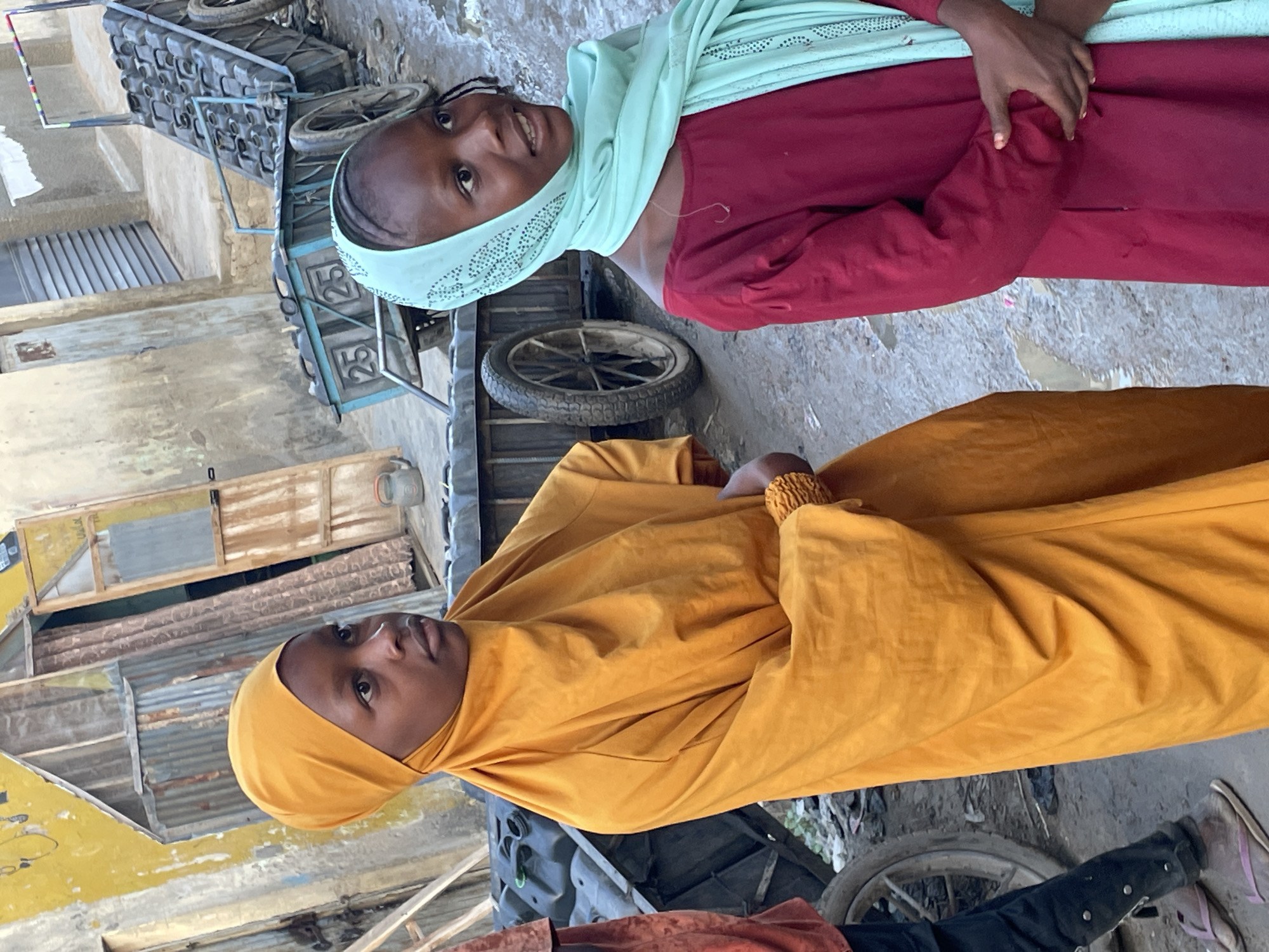 Two young girls pose outside in what appears to be an urban environment. The first girl is wearing a maroon dress and a light green headscarf, and she smiles at the camera. The second girl is wearing a yellow robe and headscarf. Various wheeled vehicles are visible in the background. The setting is outdoors.