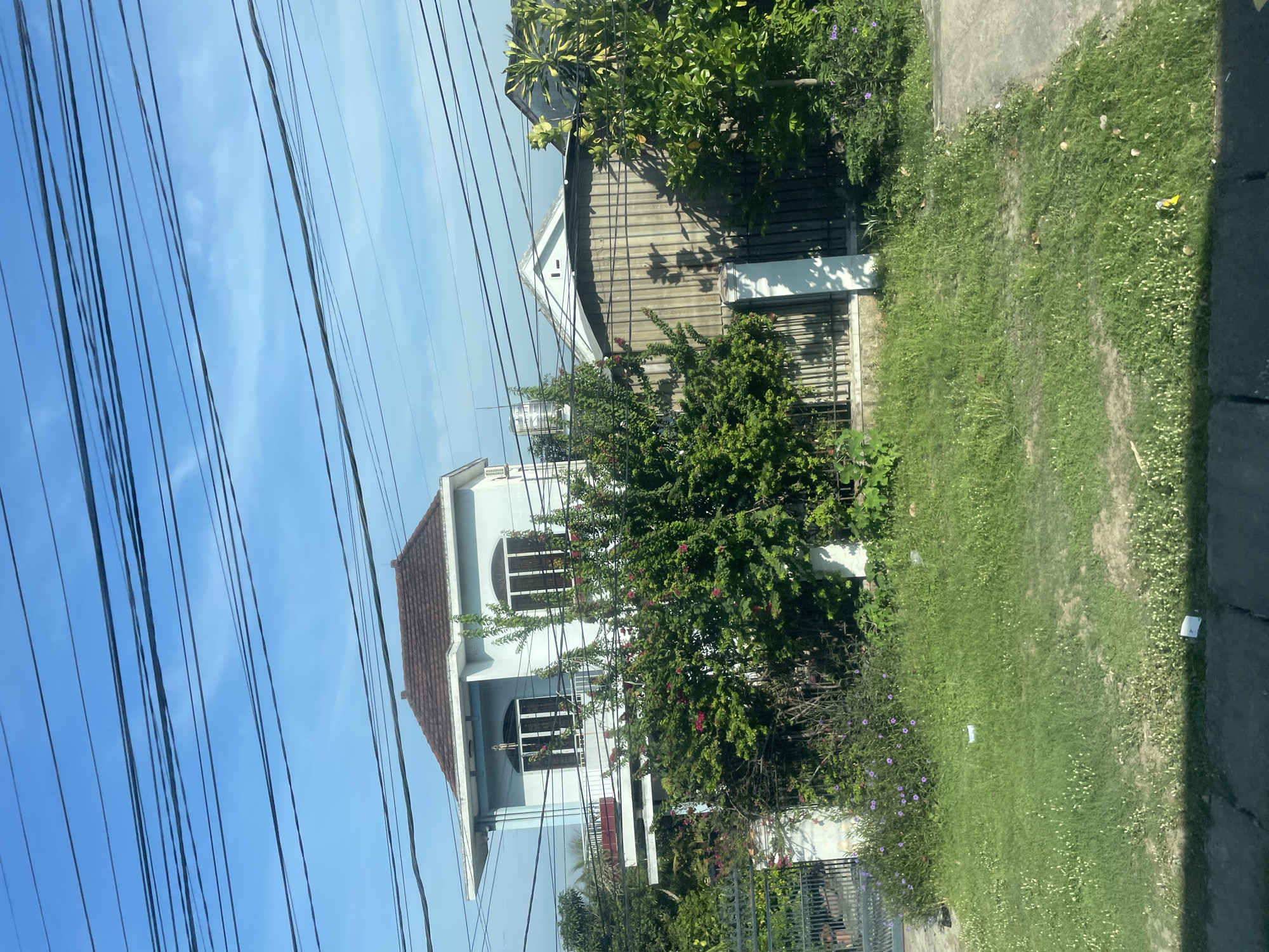 The image depicts a residential area in Suối Hiệp, Vietnam, characterized by a two-story house with a brown tiled roof and light blue walls, situated behind a fence and dense greenery. The house has balconies and arched windows on the second floor. A metal gate is visible to the left, leading to the property. The foreground features a patch of overgrown grass with scattered small white flowers and some litter. Overhead, a network of black electrical wires crisscrosses the sky, which is a clear blue. To the right of the main house, another building with a white, triangular roof and corrugated metal siding is partially visible, also behind fencing and foliage. The overall impression is of a somewhat unkempt, but lived-in, suburban environment.