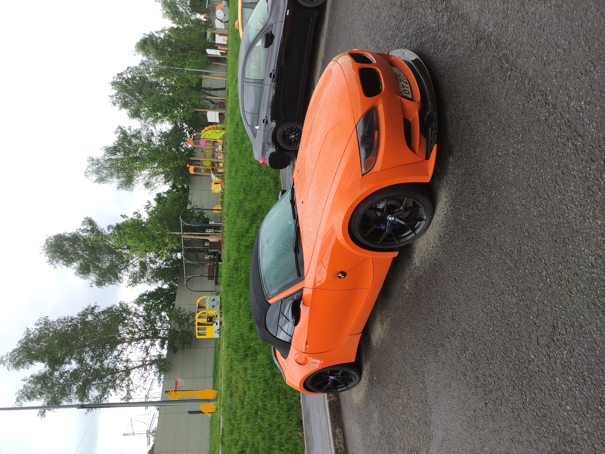 The image shows an orange BMW Z4 parked on an asphalt surface next to a curb. To the left of the car is a grassy area with several trees and a playground with yellow and orange equipment. Another dark-colored car is partially visible behind the orange car. The license plate visible on the orange car suggests the location is likely within Russia, as the license plate appears to be Russian format, in the city of Moscow, Russia.