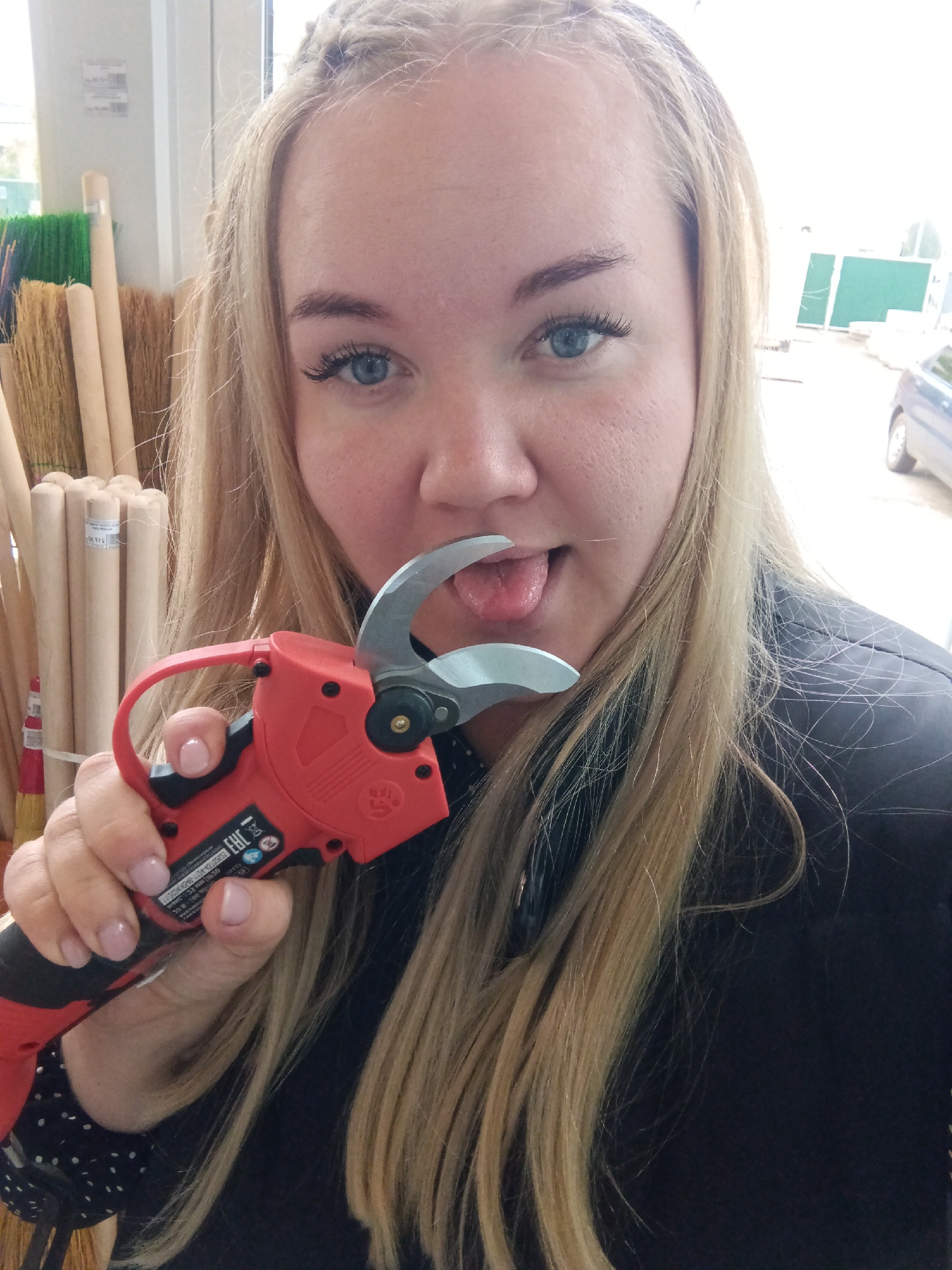 A blonde woman with blue eyes is captured in a playful pose, looking directly at the camera with her tongue sticking out. She holds a red and black electric pruning shears with open blades near her mouth. Her hair is long and blonde, with a small braid visible on her forehead, and she wears a dark top, possibly a leather jacket over a black polka-dot shirt.
The setting appears to be an indoor retail store, such as a hardware or garden supply center, with a large window looking outdoors. On the left side of the image, stacks of long, light-colored wooden handles, likely for brooms or garden tools, are visible, alongside some colorful bristles. Through the window on the right, an outdoor scene shows a paved area, green construction barriers or fencing, and part of a dark-colored car. The natural light indicates it is daytime.
Notable details include the "EAC" certification logo visible on the electric shears, along with other technical markings such as "A: 4/12V DC" and "2020.10". The woman's expression conveys a lighthearted and mischievous mood. The location context of "Foki, Russia" is consistent with the presence of the EAC certification mark on the tool.