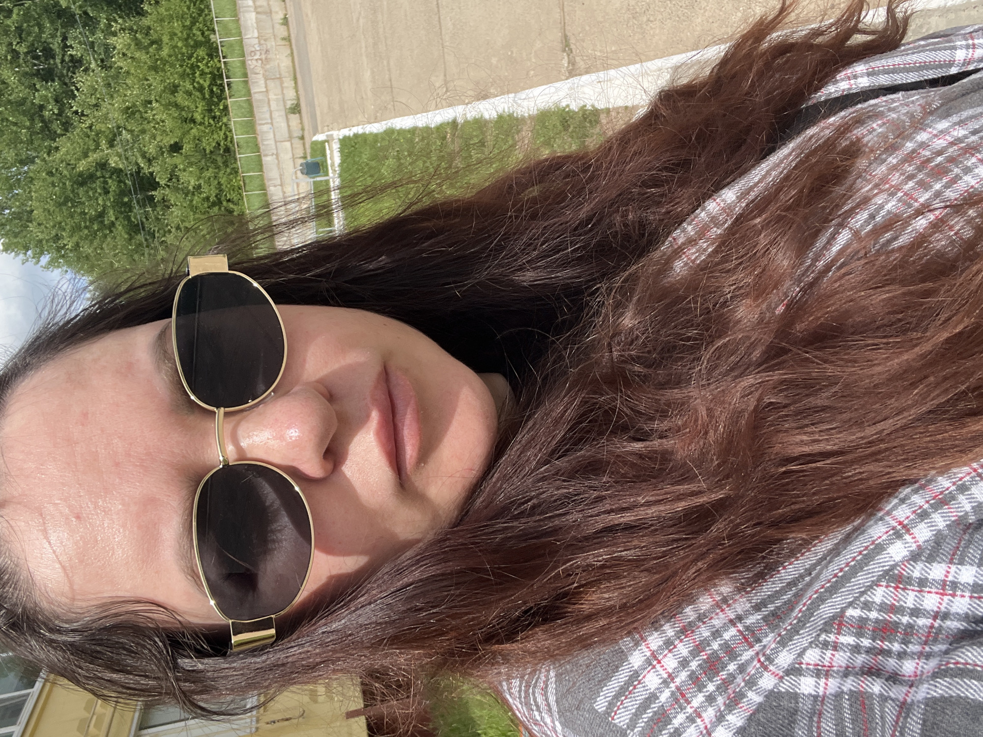 This close-up portrait, taken in Noviy, Russia, features a person, likely female, positioned outdoors. The image is rotated 90 degrees clockwise, showing the individual's face in a tilted orientation. She has long, dark brown hair that appears thick and slightly wavy. Her eyes are obscured by large, gold-framed sunglasses with dark lenses. The visible portion of her face shows a neutral expression with slightly parted lips. She is wearing a grey plaid shirt with red and white accents.
The background reveals an outdoor setting under bright daylight, indicated by strong sunlight creating highlights on her hair and skin. Lush green trees and grass are prominent, suggesting a park-like or suburban environment. Below the green area, a paved or concrete surface is visible, possibly a road or walkway, bordered by a white railing or fence. Faint outlines of buildings can be seen in the distance through the foliage, indicating an urban or suburban location. No specific activity is depicted beyond posing for the photograph. There is no visible text in the image.