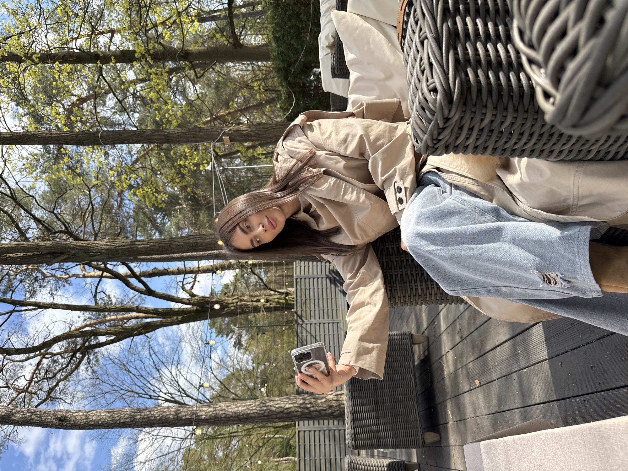 A young woman is sitting in an outdoor setting, taking a selfie with her phone. She is wearing a tan trench coat and blue jeans with a rip in the knee. The background features trees and foliage, and there are string lights visible in the trees. The woman is sitting on a wicker chair on a wooden deck. A wicker basket is partially visible in the foreground. The location is Konstancin-Jeziorna, Poland.