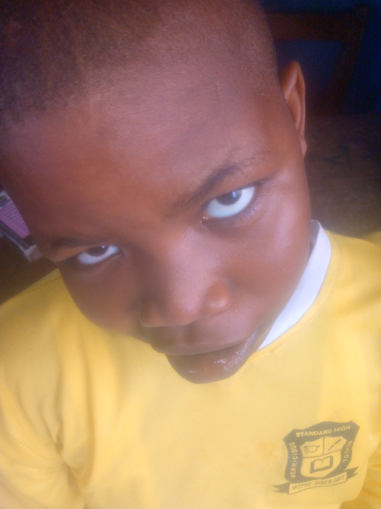 A close-up, angled shot shows a young boy with dark skin and short-cropped hair. He is wearing a yellow shirt with a white collar. On the right side of the shirt, there is a dark emblem that reads "STANDARD HIGH SCHOOL" and the motto "NOTTO GOD'S GIFT." The boy's gaze is directed upward toward the camera, with a slightly raised eyebrow. The background is blurred and indistinct, but it appears to be indoors. The location is in the city of Unknown, Unknown.