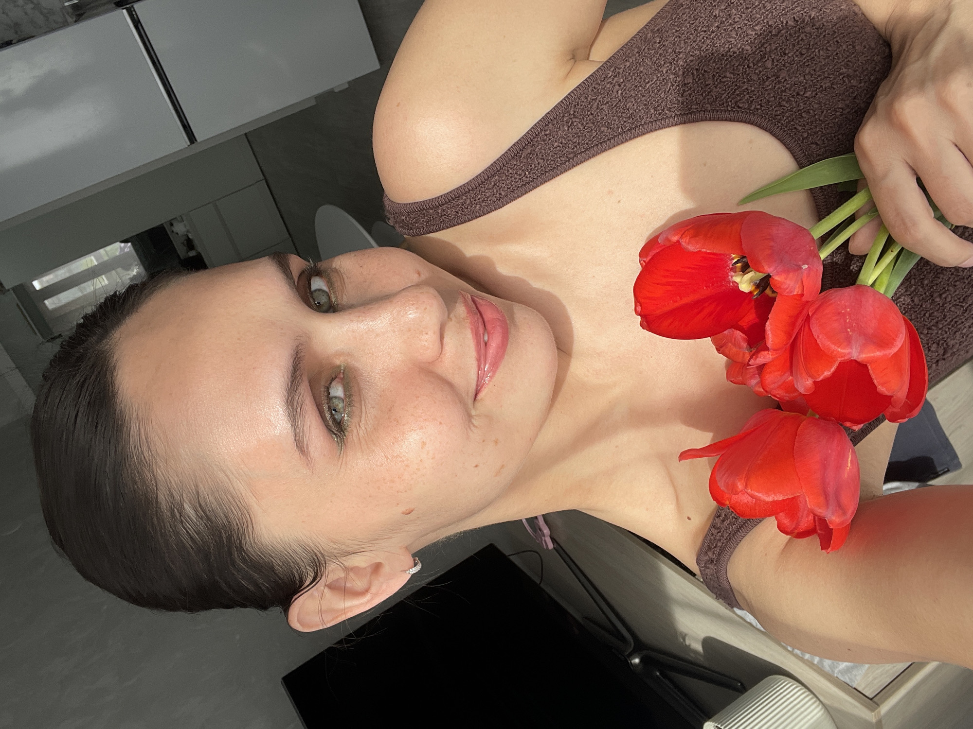 A woman is holding three red tulips in an indoor setting. She is wearing a brown tank top. The location is unknown.