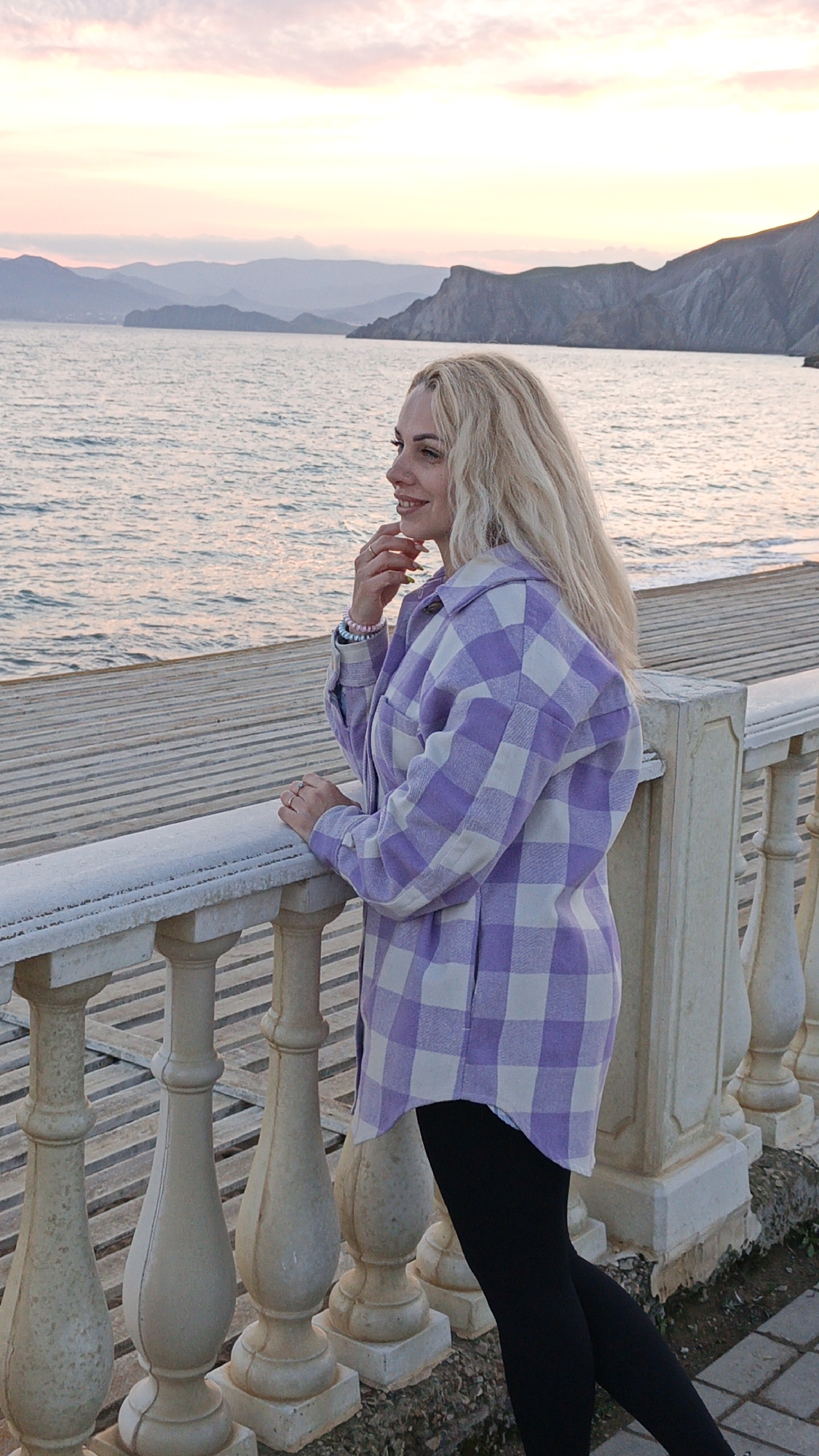 A woman is standing on a pier in Unknown, Unknown, looking out at the water. She has blonde hair and is wearing a purple and white plaid shirt and black leggings. She is resting her hand on a white railing. In the background, there are mountains and a pink and purple sky.
