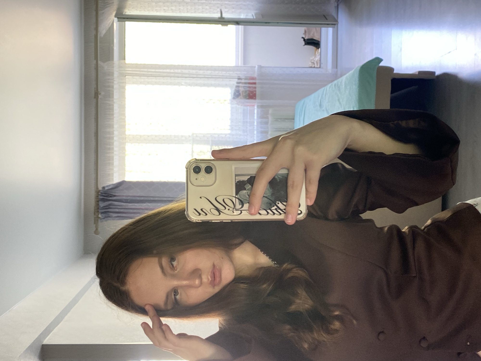 A young woman takes a selfie in a room. She holds a white cell phone up to the mirror, her hand visible holding it. The woman is facing the mirror, and her face is visible in the reflection. She has long, brown hair and is wearing a brown jacket. Behind her is a window with white curtains, a bed with a light blue blanket, and light-colored walls. It's impossible to determine the specific location in Izhevsk, Russia, without further information.