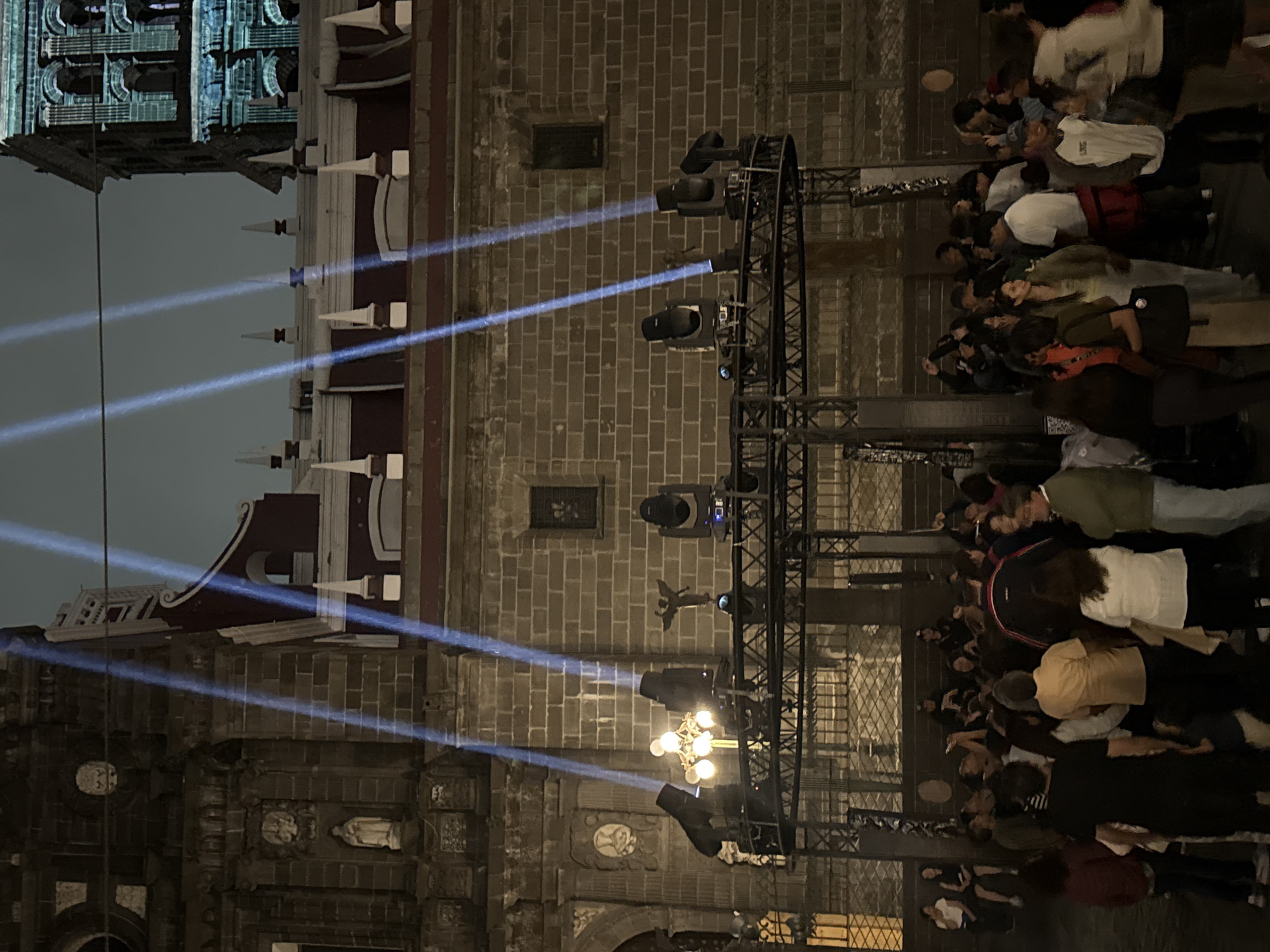 This nocturnal image, set in Puebla, Mexico, showcases a large, historic stone building illuminated by a light show, with a crowd of spectators gathered. The building, likely a colonial-era church or cathedral, features textured stone block masonry, decorative architectural elements, and white spires along its roofline. Statues are visible within niches on its facade, including a robed figure. In the upper left, another ornate, partially visible structure, perhaps a tower or dome, is illuminated with blue light.
In the foreground, a tall metal truss structure supports multiple professional stage lights, which project intense blue-white beams of light across the scene and onto the building. An antique-style street lamp with several glowing globes is also present near the base of the truss, casting a warm glow.
A sizable crowd of people occupies the right side of the frame, positioned to view the illuminated building and the light projections. They are casually dressed, mostly seen from behind or in profile, engrossed in the spectacle. The dark sky confirms the nighttime setting, and the distinct light beams indicate clear weather conditions. The scene suggests a public event or cultural display where light art is projected onto historical architecture. No discernible text is visible in the image.
