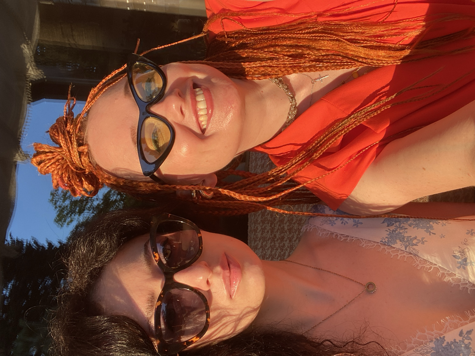 A close-up outdoor portrait features two young women, illuminated by warm, golden hour sunlight in the city of Sochi, Russia.
On the left, a woman with dark, curly hair wears tortoiseshell cat-eye sunglasses, partially obscuring her eyes. Her lips are a soft pink. She wears a light-colored dress with blue floral patterns and white lace trim, adorned with a thin gold chain necklace featuring a circular pendant. Her expression is neutral as she looks towards the camera.
On the right, a woman with vibrant orange micro-braids, partially styled into a high bun, grins broadly, revealing her teeth. She wears black cat-eye sunglasses with yellow-tinted lenses, and her skin appears slightly dewy. She is dressed in a bright orange collared top and wears multiple necklaces: a chunky, sparkling choker and a longer silver chain with a crucifix pendant. A decorative ear cuff is visible on her right ear.
The intense, low-angle sunlight casts prominent shadows and highlights across both individuals. The background shows blurred natural elements, including dark green trees against a clear blue sky on the left, and indistinct darker structures or interior elements on the right, suggesting an urban or semi-urban outdoor setting, possibly a balcony or terrace.