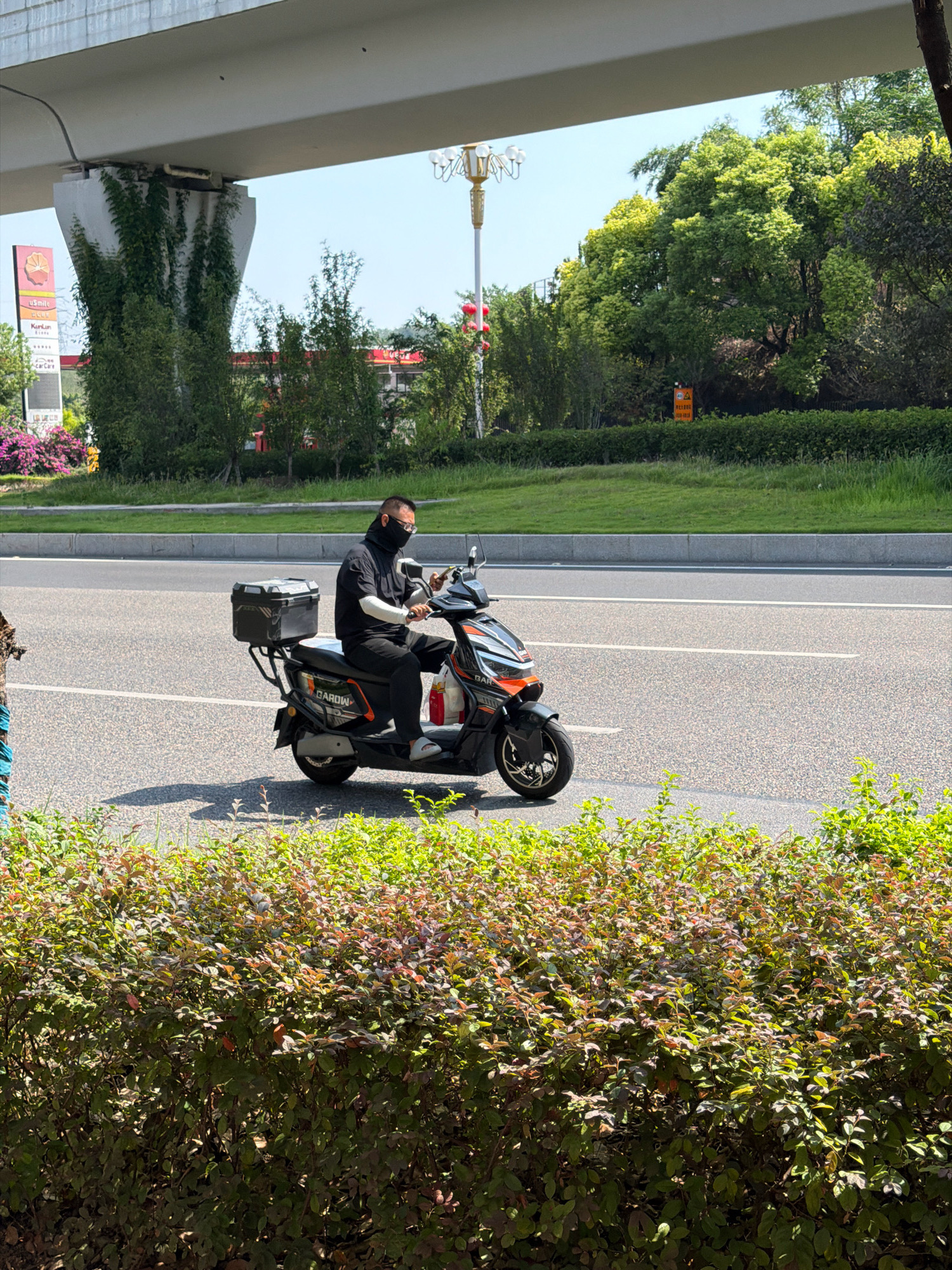 In Jiangjin, China, a man wearing a black face mask and white arm coverings rides a black and orange electric scooter on a paved road. The scooter has a black storage box mounted on the rear. The man is holding a mobile phone in his right hand while riding. The scene is framed by a low, dense hedge in the foreground and a concrete overpass with overgrown vines in the background. Trees and a street lamp are visible beyond the road, with a clear sky above.