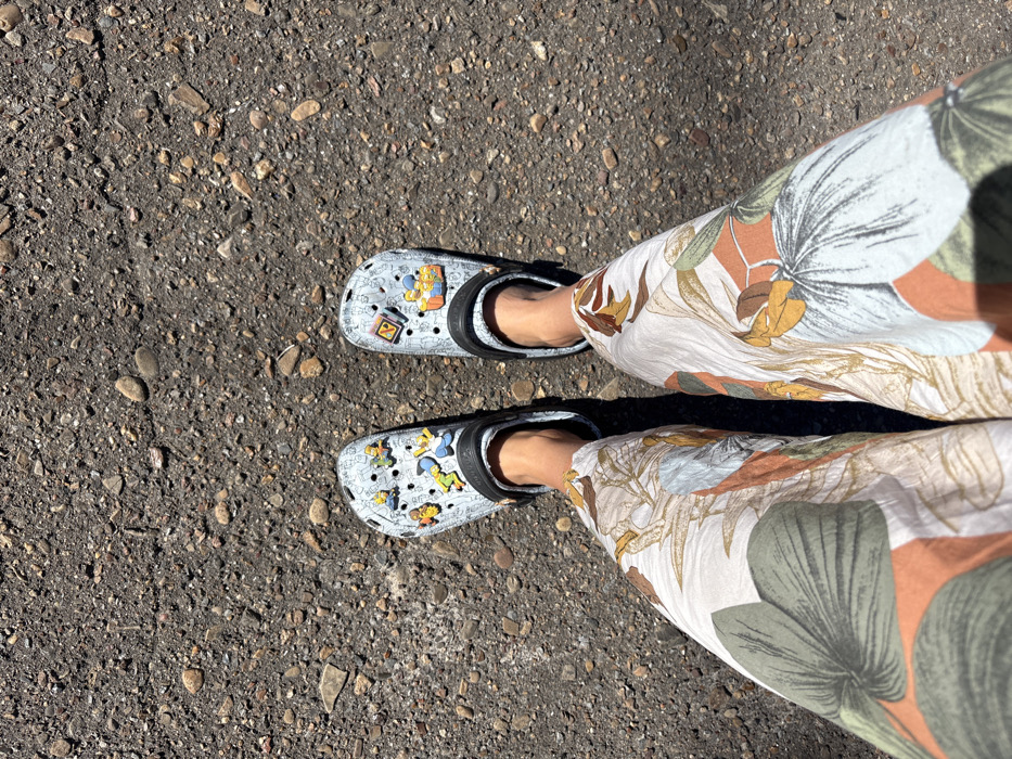 An overhead view captures the lower legs and feet of an individual standing on a dark, granular ground surface in Kropachyovo, Russia. The person wears wide-leg trousers featuring a botanical print with large green leaves and abstract orange and brown shapes against a light background. On their bare feet are light grey Crocs-style clogs with black straps. The left clog is adorned with multiple Jibbitz-style charms, predominantly depicting characters from "The Simpsons," including Bart, Lisa, Homer, and Marge. The right clog displays charms including an "N" block and a depiction of "The Simpsons" family seated on a couch. The ground beneath the individual is dark grey, textured asphalt or packed gravel, embedded with numerous small stones and pebbles. Shadows are visible, suggesting bright, direct overhead lighting.