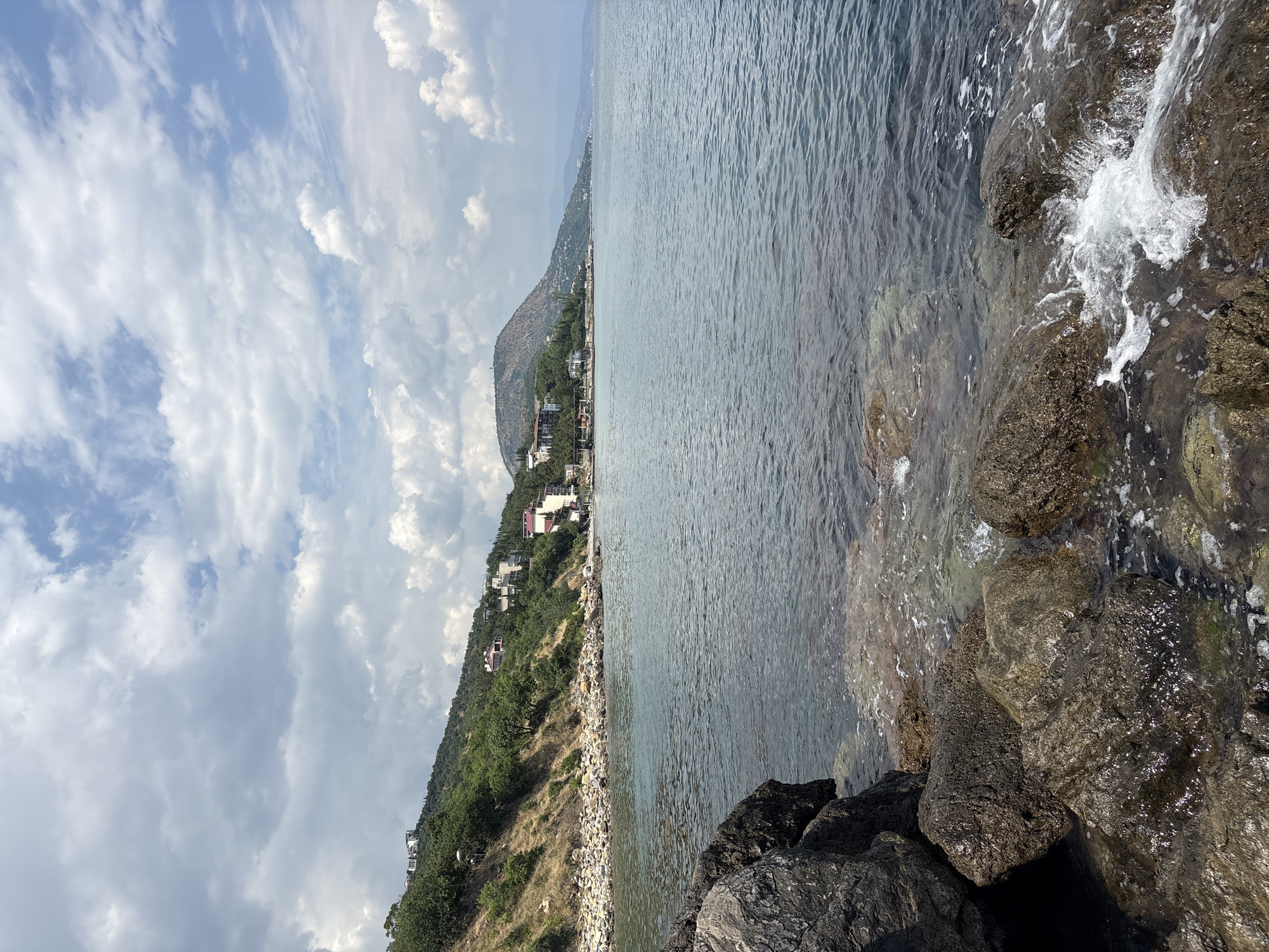The image depicts a coastal scene with a rocky shoreline in the foreground. The sea is calm with ripples on the surface and appears to be a clear blue-green color. In the middle ground, a rocky beach leads up to a vegetated hillside dotted with houses. The houses are modern in design, with white and red facades, and are spread out along the slope. Further in the distance, a larger, rounded hill is visible, and beyond that, the coastline recedes into the horizon. The sky is partly cloudy with dramatic white and gray clouds against a light blue background. The overall impression is a sunny day at a Mediterranean-style coastal town.
