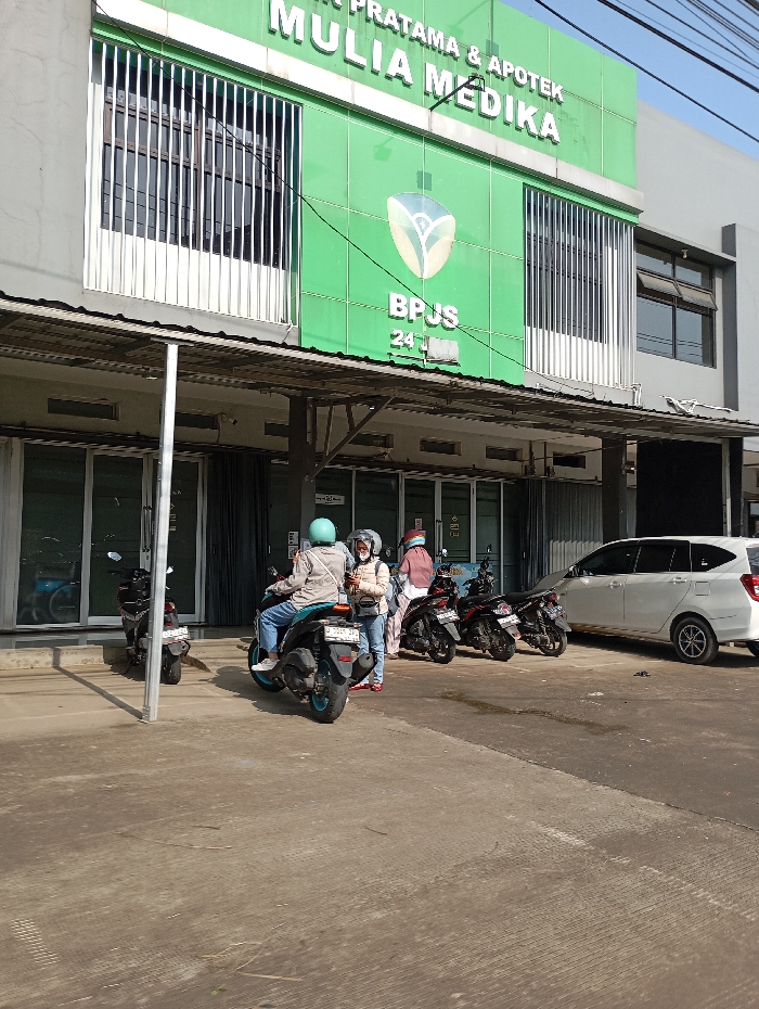 The image depicts the exterior of Klinik Pratama & Apotek Mulia Medika, a medical clinic and pharmacy, located in Pataruman, Indonesia, under clear daylight conditions. The two-story building features a prominent green facade on its upper level, displaying "KLINIK PRATAMA & APOTEK MULIA MEDIKA" and "BPJS 24H" branding. Windows on the upper floor are protected by vertical metal bars. A gray, corrugated awning extends over the ground floor entrance, supported by metal poles.
On the concrete forecourt in front of the clinic, four individuals are present. One person, wearing a light-colored jacket, blue jeans, and a light blue helmet, is seated on a dark scooter with teal-colored wheels, facing right. Another person, dressed in a light jacket, blue jeans, a white helmet, and a face mask, stands beside this scooter, holding a phone. Two other individuals stand further right near several parked dark motorcycles. A white multi-purpose vehicle is partially visible, parked on the far right of the scene. Multiple scooters are parked on the paved area, suggesting a common mode of transport for visitors.