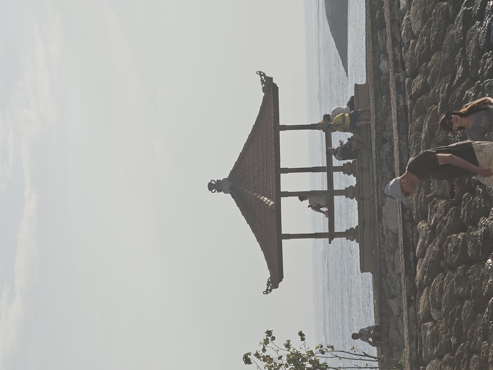 The image depicts a scene at the beachfront in Denpasar, Indonesia. In the foreground, a man wearing a dark t-shirt and a cap is partially visible, facing towards the right of the frame. Beside him, a woman with sunglasses and a maroon baseball cap is looking towards the left. They are standing on a rocky shoreline.
In the midground, a traditional Balinese gazebo with a reddish-brown tiled roof stands near the water. Several individuals are seated on the ledge of the gazebo. To the left of the gazebo, a young person is sitting alone on the stone embankment, facing the ocean. Further to the left, a small tree with sparse green leaves grows.
The background features the ocean with gentle waves and a hazy sky. The overall atmosphere appears serene, possibly during a late afternoon or early morning due to the soft lighting. The location suggests a popular tourist spot or a local gathering place along the coast.