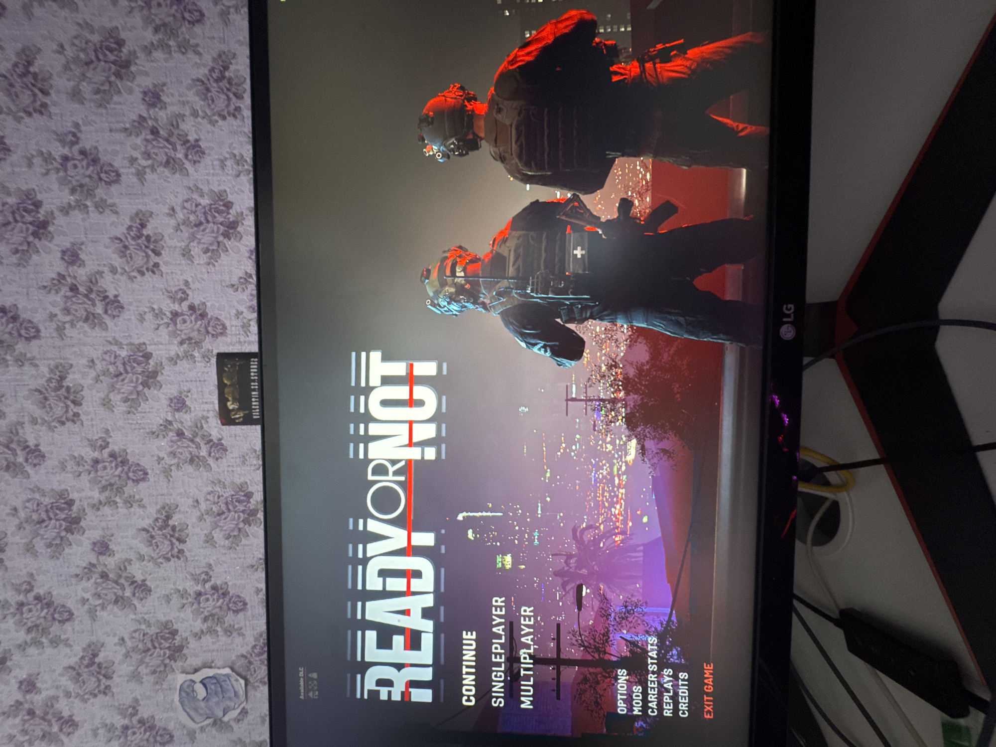 This media file captures the title screen of the video game "Ready or Not," displayed on a monitor. The screen features two heavily armed individuals in tactical gear, silhouetted against a vibrant cityscape at night. The city skyline is illuminated with countless lights, suggesting a sprawling urban environment. The overall mood is intense and action-oriented, consistent with a tactical shooter game.
The game's title, "READY OR NOT," is prominently displayed in large, bold, white lettering with a red line striking through the "O." Below the title, a menu of options is visible: "CONTINUE," "SINGLEPLAYER," "MULTIPLAYER," "OPTIONS," "MODS," "CAREER STATS," "REPLAYS," "CREDITS," and "EXIT GAME." The background image depicts two figures in tactical gear, one slightly behind the other, both facing towards the viewer. They are equipped with helmets, vests, and what appear to be firearms. The lighting is dramatic, with red hues casting an ominous glow on the figures and the cityscape.
The monitor is positioned against a wall with floral wallpaper in shades of purple and white. A small, dark object, possibly a sticker or a small picture, is adhered to the wallpaper on the left side of the frame. Cables and other electronic equipment are visible to the right of the monitor, suggesting a home gaming setup. The presence of an LG logo on the monitor's bezel indicates the brand of the display. The provided location context of Zolotonosha, Ukraine, is external information and not directly discernible from the image itself.