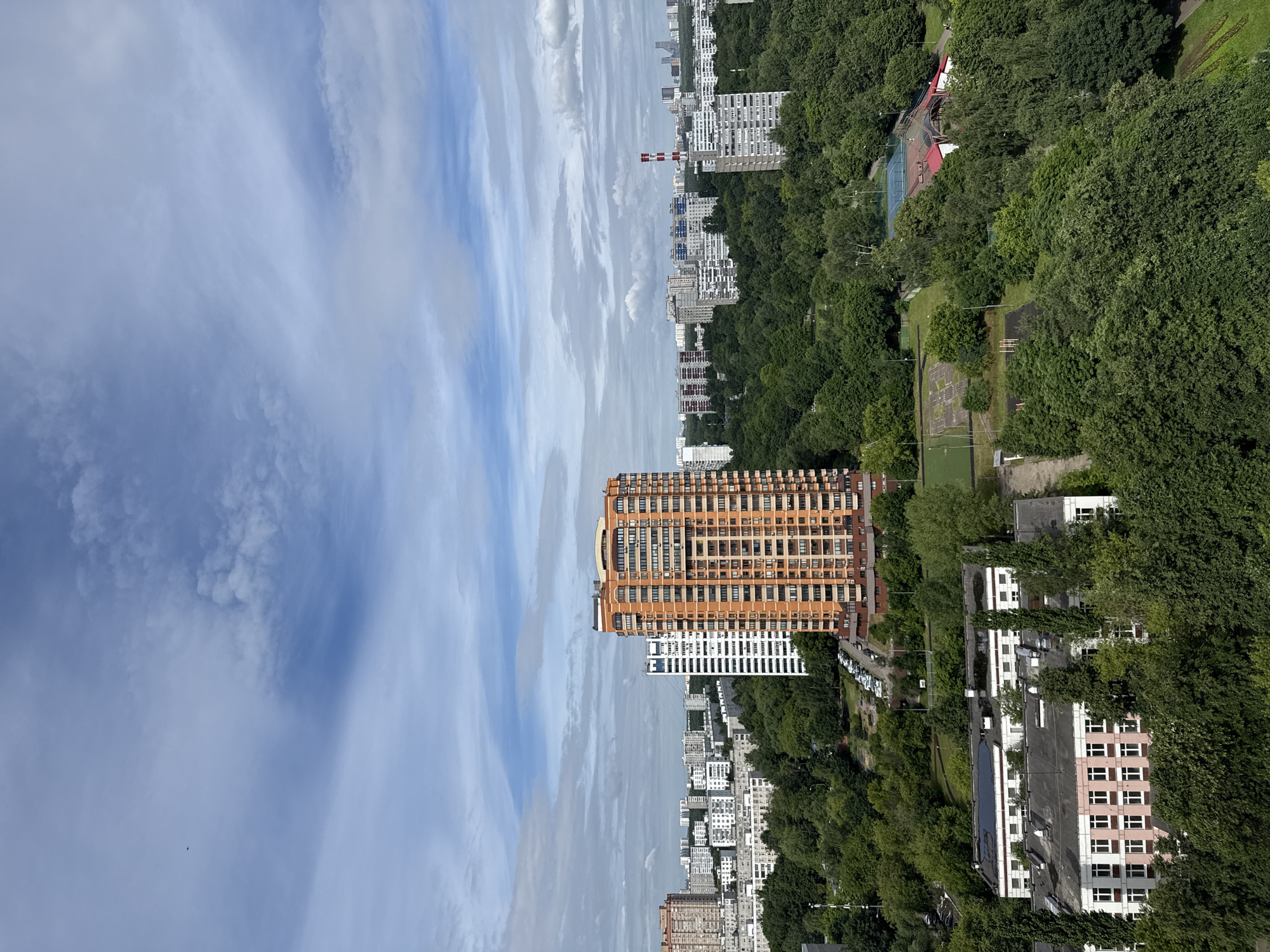 An aerial shot captures a cityscape in Moscow, Russia, dominated by an overcast sky with scattered white clouds. In the foreground, lush green trees form a dense canopy, partially obscuring several buildings. To the left, a low-rise building with a pink and white facade is visible. Further back, a cluster of multi-story residential buildings, varying in architectural style and color, punctuate the urban landscape. A prominent, tall, orange-colored apartment building stands out in the mid-ground. In the background, a dense expanse of trees separates the nearer buildings from a more distant array of high-rise structures. A single small bird is discernible in the upper left portion of the sky. The scene suggests a residential area with ample green spaces.