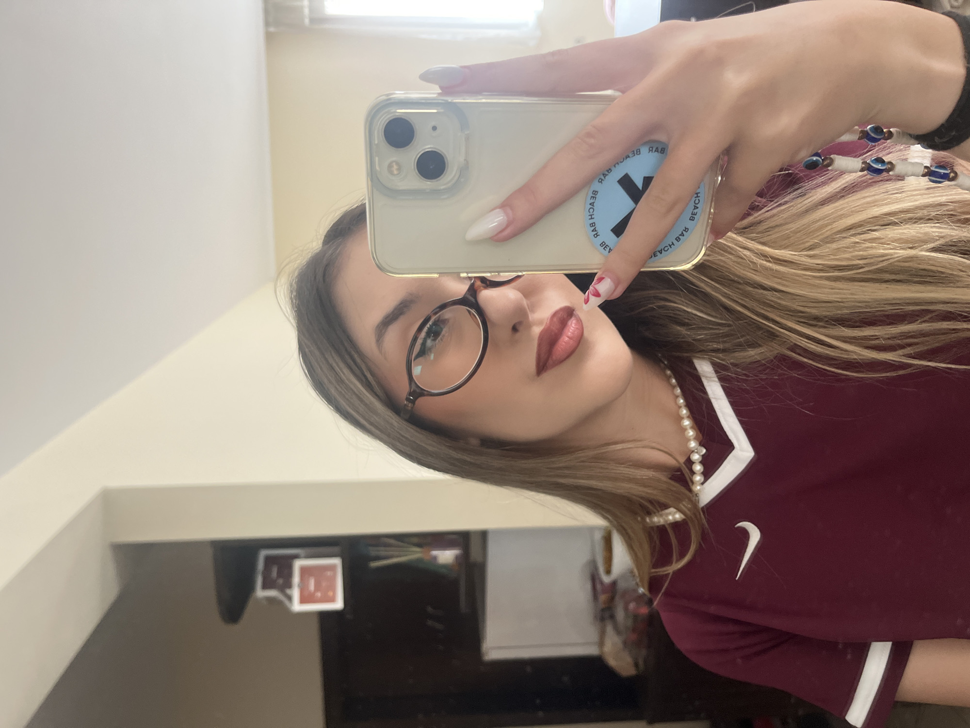 A young woman is captured in a mirror selfie, seen from the chest up, with her head tilted to the right. She is holding a light-colored smartphone, likely an iPhone, in her right hand, positioned to take the photograph. Her long, light brown hair frames her face. She wears round, dark-framed glasses and has dark reddish lipstick.
Her attire includes a maroon or dark red collared top with white trim along the neckline and sleeves, featuring a white Nike swoosh logo on the chest. A white pearl necklace is visible around her neck. Her fingernails are long, pointed, and painted white, with a subtle floral design on her right pinky finger. A bracelet adorned with blue evil eye beads is on her right wrist.
The smartphone's clear case reveals a round, light blue sticker on its back, which displays a large white 'X' and the mirrored text "BEACH BAR" repeated around its perimeter.
The background, reflected in the mirror, reveals a simple indoor setting. Light-colored, possibly off-white or cream, walls are visible, and in the lower part of the reflection, there appears to be dark furniture or shelving. The lighting suggests an indoor environment. The image is taken in Sofia, Bulgaria.