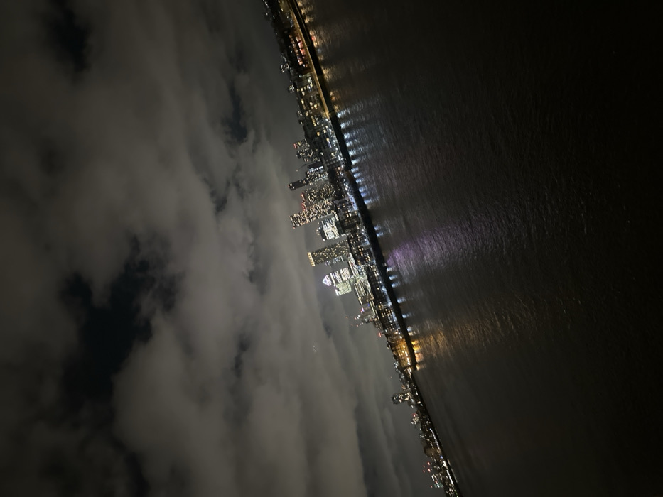 An oblique nighttime cityscape reveals a dense cluster of illuminated skyscrapers, their lights reflecting on the adjacent dark water. Clouds partially obscure the lower portion of the city, creating a misty or foggy atmosphere. Lights stretch along the shoreline, marking a clear division between the city and the water. The angle of the image creates a unique perspective, emphasizing the verticality of the buildings and the vastness of the night.