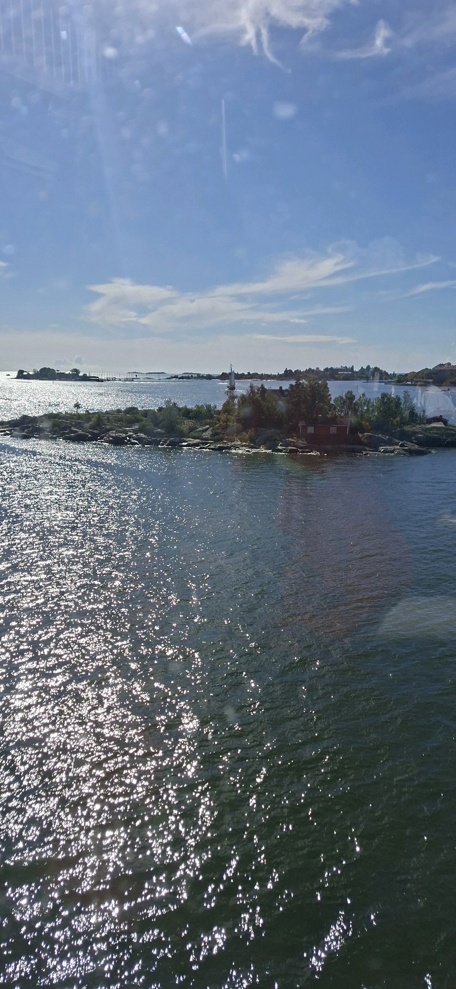 The image depicts a sunny day with a clear blue sky and scattered clouds, over a body of water with islands and boats. The water is dark blue and reflects the sunlight, creating a shimmering effect. In the foreground, the water is closer and more detailed, showing the light refractions. Several rocky islands are visible, dotted with trees and some small buildings. One prominent island in the mid-ground has a small red building with a dark roof, and a few trees. Further in the distance, more islands and the coastline are visible. There are several boats on the water; a sailboat with white sails is moving to the right in the mid-ground. The scene appears to be a picturesque view of the Helsinki archipelago, likely from a ferry or boat, showcasing the natural beauty and maritime character of the area.