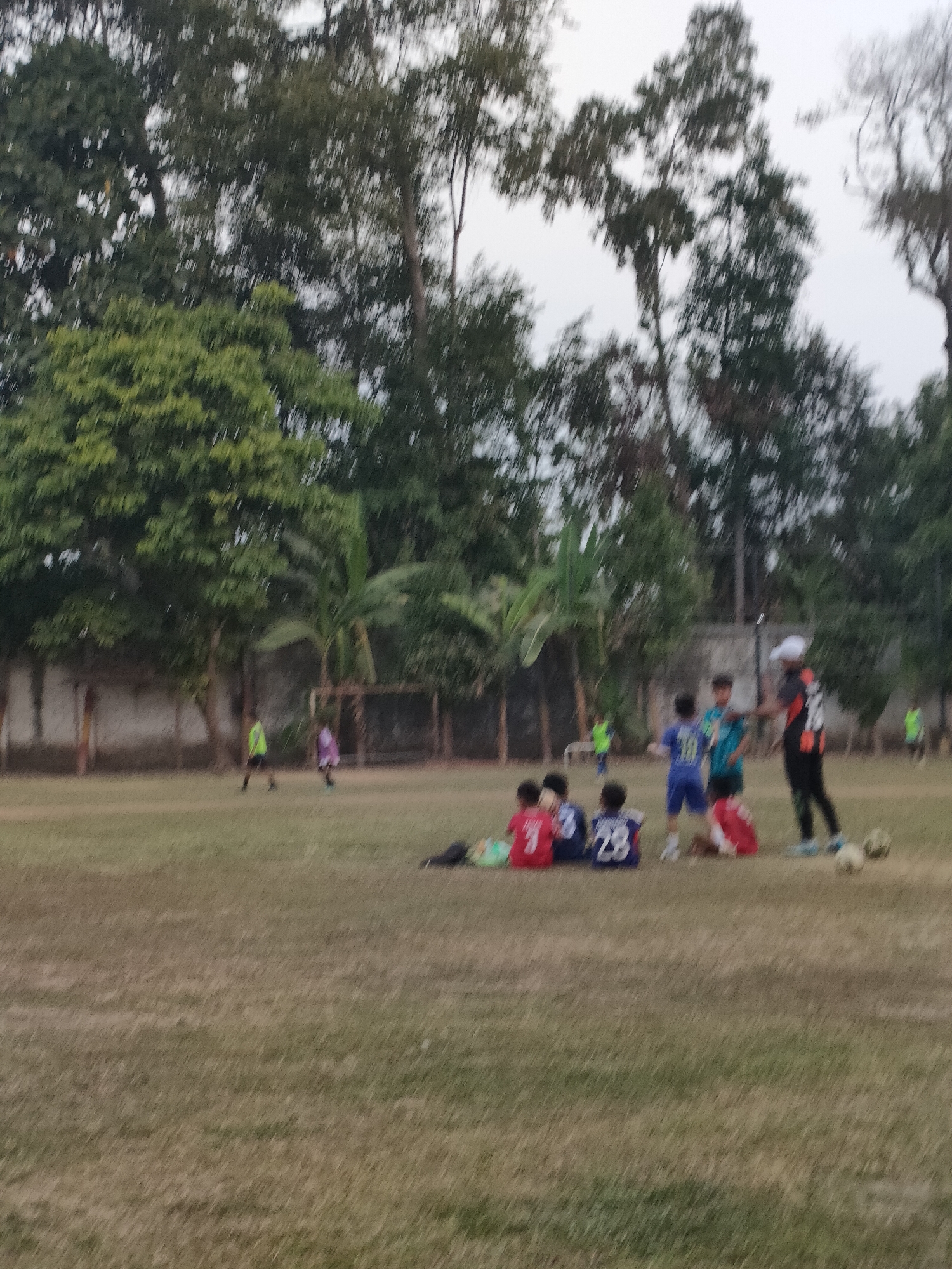 The image depicts an outdoor scene at a grassy sports field, possibly a soccer field, in Banguntapan, Indonesia. In the foreground, several children are seated on the grass, some wearing soccer jerseys. Behind them, a group of individuals, including children and an adult wearing a white cap and an orange and black jersey, appear to be engaged in a soccer-related activity, possibly a practice or training session. Two children in soccer uniforms are standing and interacting with the adult. In the background, trees line the edge of the field, and a fence is visible. Two individuals in soccer attire can be seen further in the distance on the field, and what appear to be soccer goals are present. The overall lighting suggests it is either early morning or late afternoon.