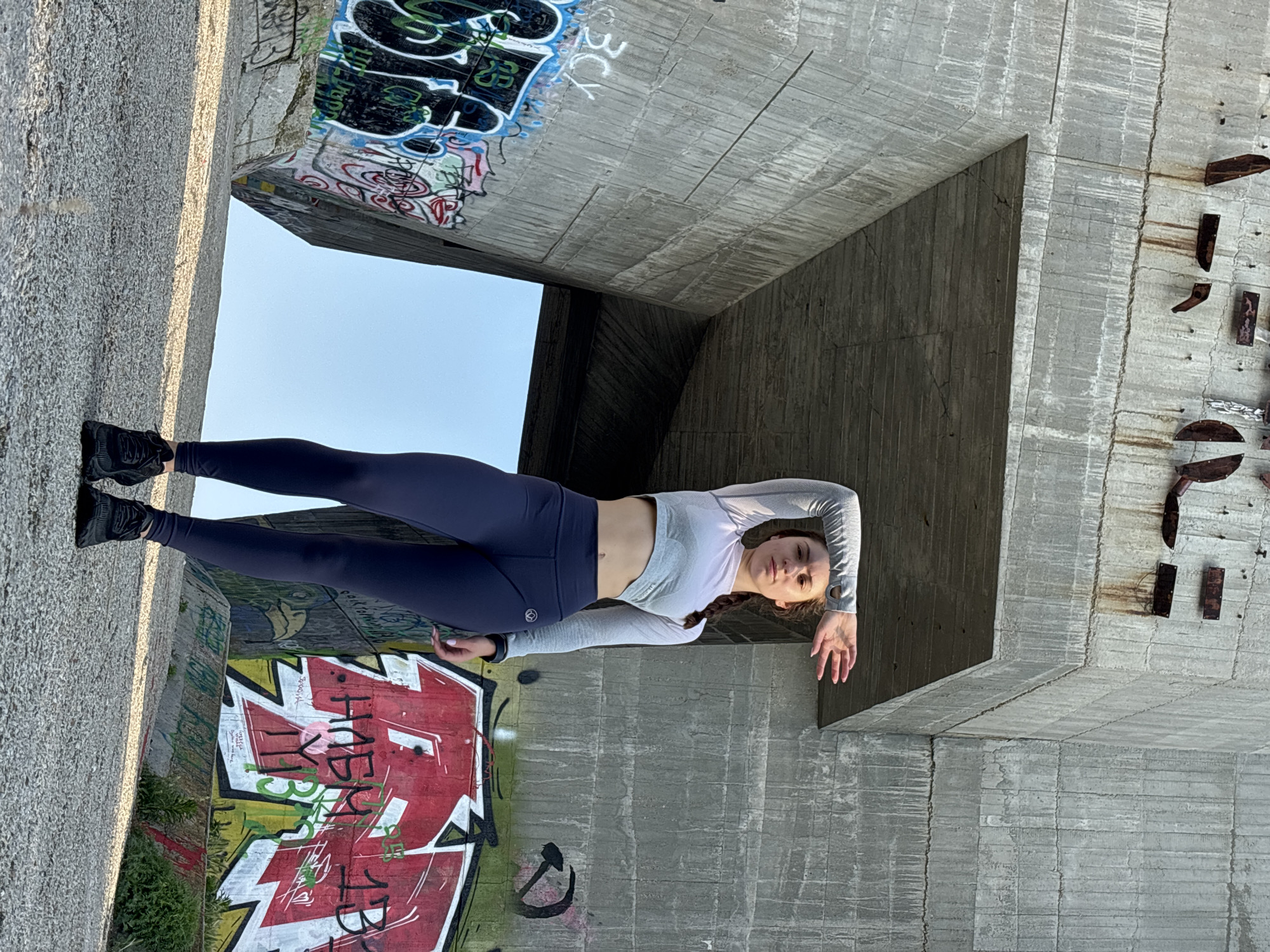 A young woman is prominently featured in the center of the frame, posing amidst a brutalist concrete structure in Varna, Bulgaria. She appears to be in her 20s or early 30s, with dark hair, possibly in a braid, visible over her left shoulder. She is dressed in athletic wear, consisting of dark navy leggings and a light grey long-sleeved crop top that exposes her midriff. Her feet are clad in black athletic shoes. Her expression is neutral as she looks directly at the camera, with her left arm extended forward and slightly bent, and her right hand resting near her hip.
The setting is characterized by massive concrete walls and an overhead structure, possibly the underside of a bridge or an architectural underpass. A large opening above the woman reveals a clear blue sky, indicating an outdoor daytime scene. The concrete surfaces show distinctive textures from formwork, as well as signs of wear, including cracks and rust stains from old metal fixtures visible on the right wall.
The environment is heavily adorned with graffiti. On the concrete wall to the woman's left, a large, vibrant graffiti piece in red, white, and green features stylized letters, with "HEB?Y" being partially discernible. Other smaller, less legible graffiti tags and drawings in various colors (green, black, white) are scattered across different concrete surfaces, including the overhead structure. A small, stylized circular logo is visible on the upper left thigh of her leggings. The lighting suggests a clear day, with soft, ambient light illuminating the scene, likely morning or late afternoon. The overall impression is a modern, urban setting with a strong artistic undercurrent due to the graffiti.