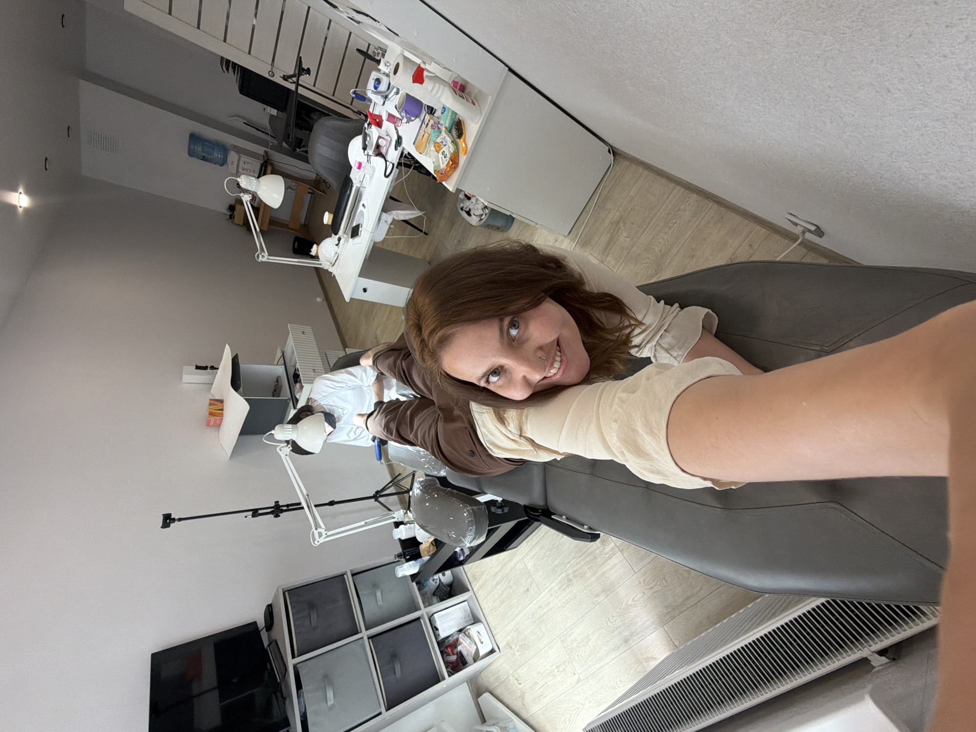 A woman is lying on a massage table in a brightly lit room, possibly a beauty salon in Izhevsk, Russia. She is looking at the camera and smiling. She is wearing a light-colored long-sleeved shirt and brown pants. Her arm is extended towards the camera, and she is wearing a beige sleeve or glove. In the background, another person in a white long-sleeved shirt and a cap is seated at a table, appearing to be working on something. There are various pieces of equipment and supplies around the room, including lamps, storage units, a television, and what appears to be a water cooler. The floor is light-colored wood.