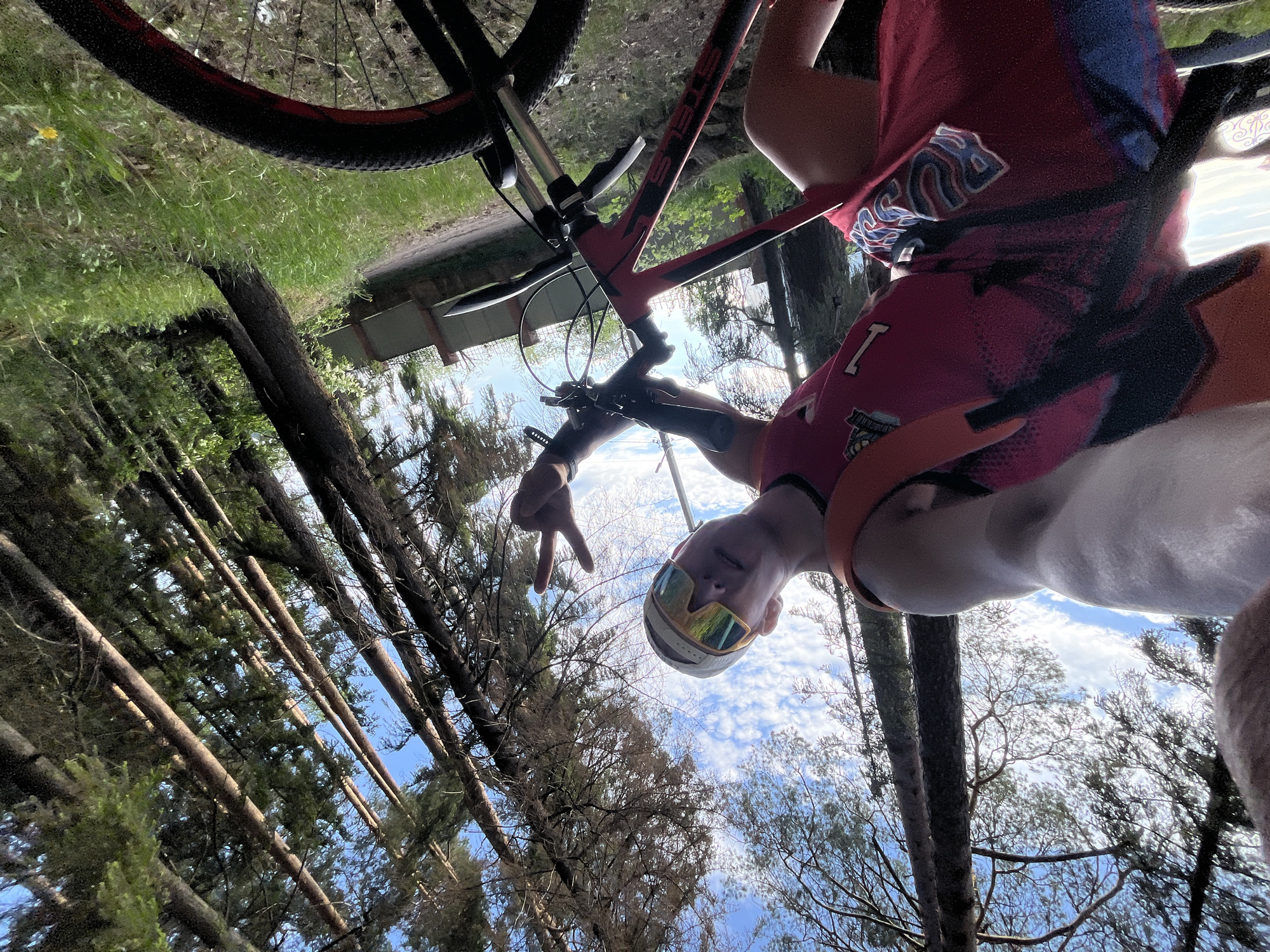 A man wearing a helmet, sunglasses, and a cycling jersey is shown in the image. He is seated on a red and black mountain bike. His left hand is raised, forming a peace sign. The background features a forest with tall trees and a blue sky with scattered clouds. A partial view of a structure with a roof is visible behind the trees. The location may be in or near the city of Chaikovsky, Russia, based on additional context. The image appears to be a selfie taken by the man while he is riding the bike.