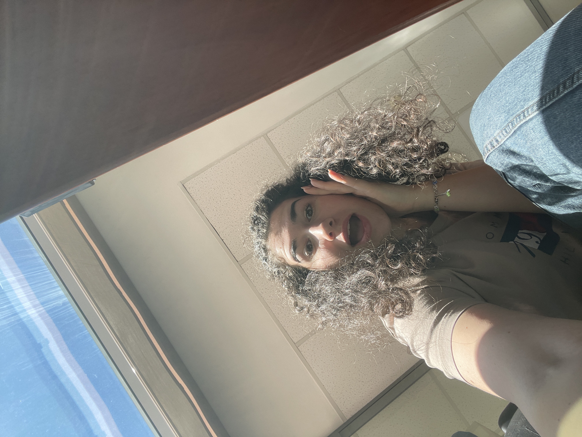 A young woman with curly hair and a surprised expression looks down at the camera. Her right hand is raised to her forehead. She is wearing a tan t-shirt and blue jeans. The photo appears to have been taken indoors, near a window with blue light. The background consists of a white tiled ceiling and a dark brown table surface. The exact location in Sofia, Bulgaria, cannot be determined from the image.