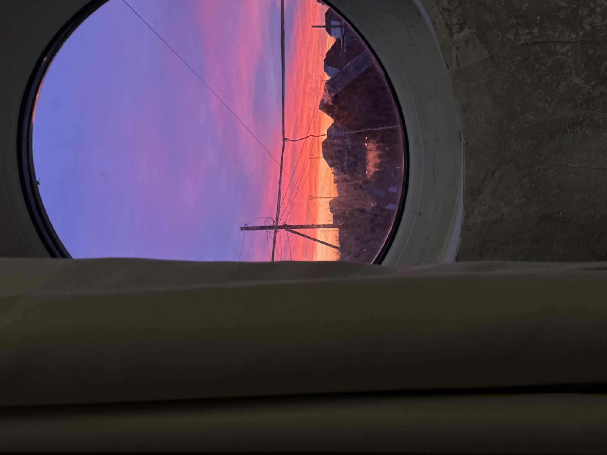The image shows a sunset as viewed through a circular window. The sky transitions from purple to pink to orange as the sun dips below the horizon. Utility poles and wires are visible silhouetted against the sky. The window frame is white. The background behind the window frame appears to be a wall, possibly made of stone or concrete. In the foreground is a section of a cream-colored curtain. The location within the city of Noviy, Russia cannot be determined from the image alone.