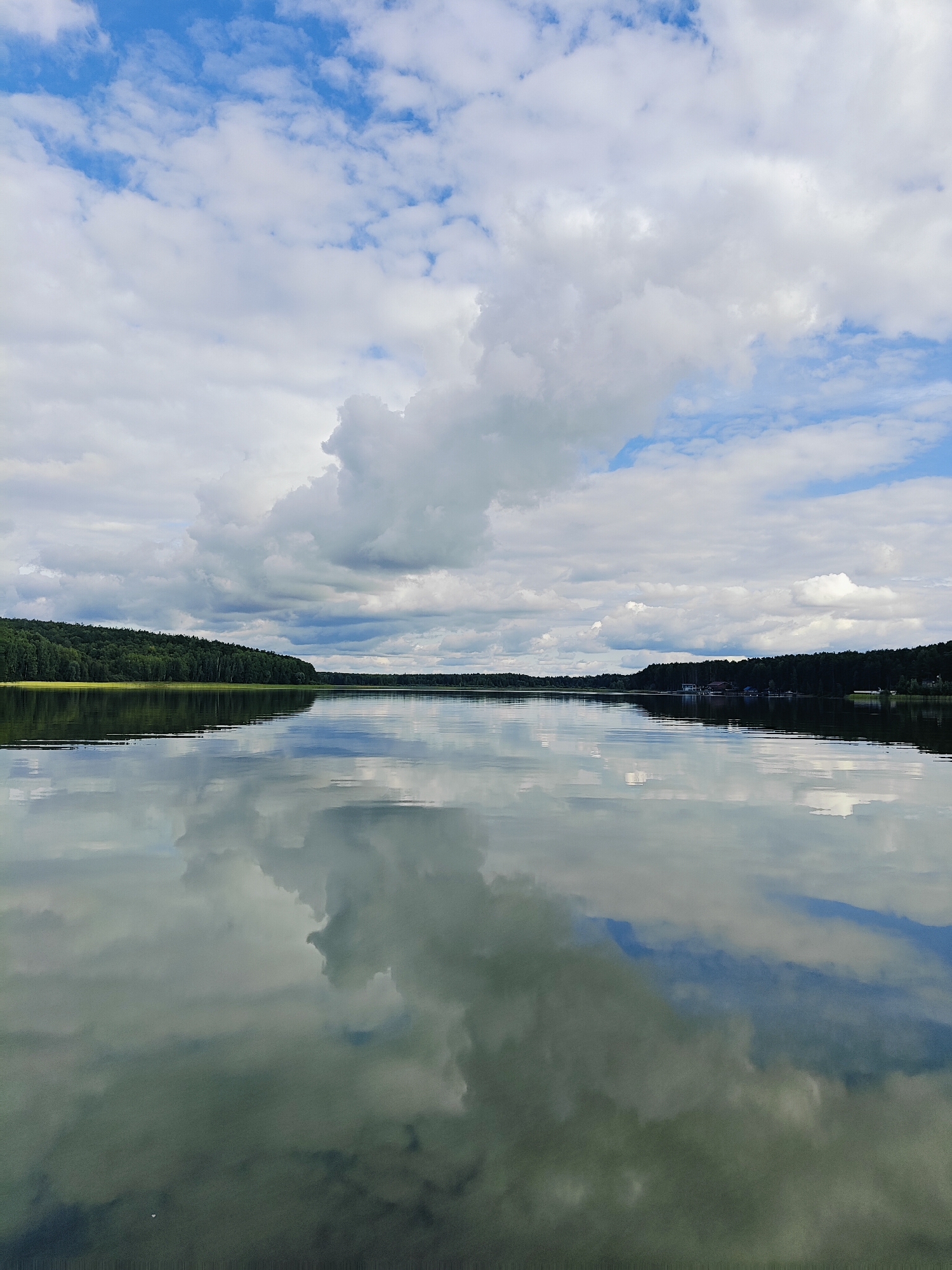 A wide shot captures a serene lake with a tree-lined shore under a dramatic sky. The water's surface acts as a mirror, reflecting the fluffy white clouds and the dark green trees with perfect clarity. The sky is a mosaic of blues and whites, with large cumulus clouds dominating the upper portion of the frame. On the left side of the image, a dense forest covers the rolling hills that lead to the lake. The right side of the lake also features a tree-lined shore with a few small buildings or docks visible in the distance. The overall impression is one of tranquility and natural beauty, suggesting a peaceful natural setting. This image is likely taken in or near the city of Chaykovsky, Russia, known for its numerous lakes and natural landscapes.