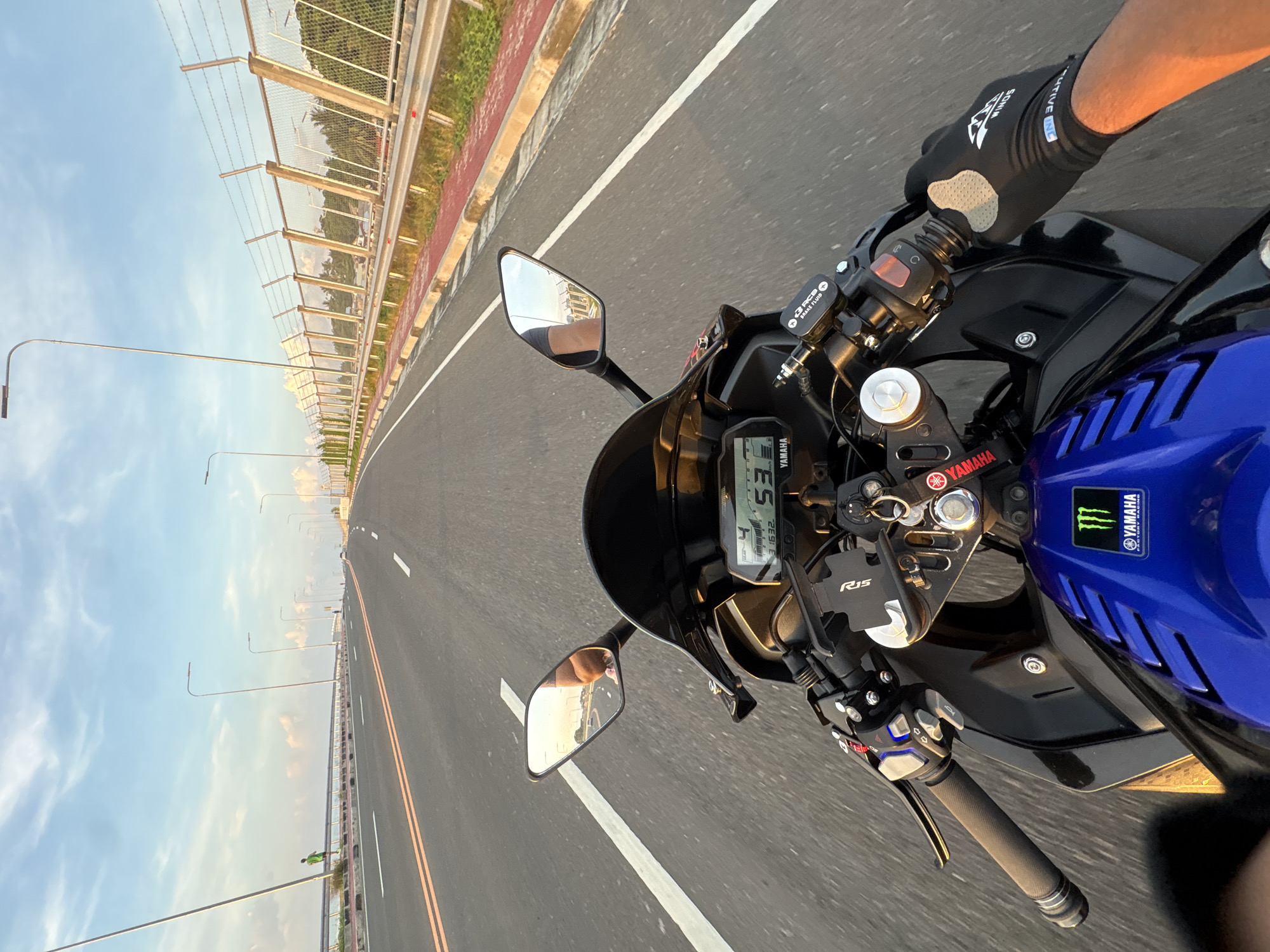A first-person perspective shot captures a motorcycle rider's view as they travel on a multi-lane paved road with double yellow lines and white lane dividers. The sky is blue with scattered white clouds, and streetlights line the right side of the road. A metal fence with barbed wire is visible beyond the streetlights, separating the road from a grassy area with some trees. In the distance, other vehicles can be seen. The motorcycle's dashboard shows a speed of 53 and a gear of 4. The rider's hands, wearing black gloves, are visible on the handlebars. The motorcycle is blue and black with "YAMAHA" branding and a Monster Energy logo on the fuel tank. The rider appears to be on the Davao Coastal Road in Davao City, Philippines, likely near the soon-to-be-completedității Panguil Bay Bridge, given the construction and infrastructure.