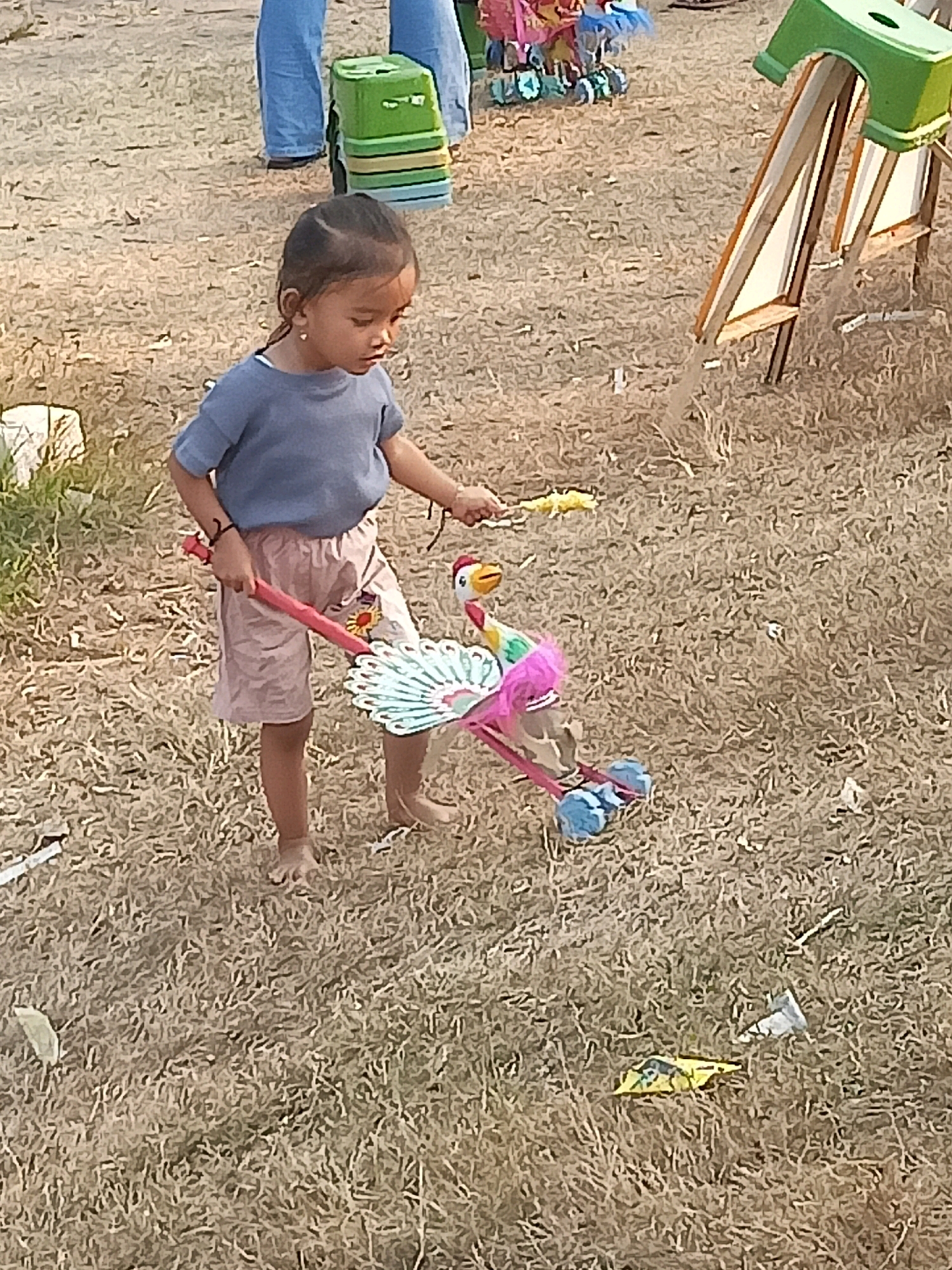A young child, likely female, is standing on dry, patchy grass in Sambiduwur, Indonesia. She is wearing a grey t-shirt and light pink shorts. The child is holding a red pole with a toy peacock lantern attached to it. The lantern has colorful decorations, including blue wheels at the bottom. The child appears to be looking down at the toy. In the background, there are parts of other people, a green trash can, and some wooden structures, suggesting an outdoor event or gathering. The ground is covered in dry grass and some scattered debris.
