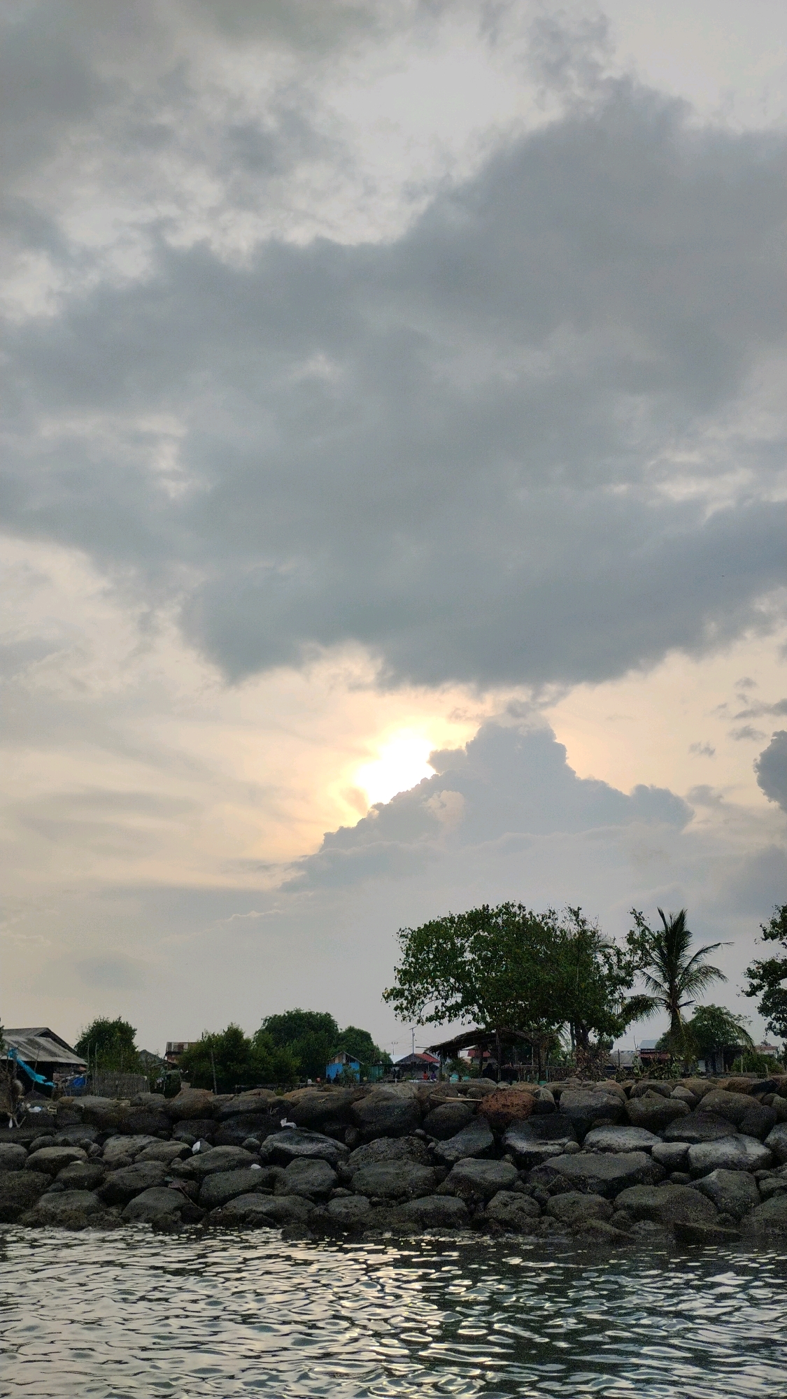 A vertical, medium shot captures a cloudy sky with the sun peeking through the clouds, casting a bright glow. Below the sky, a rocky shoreline is visible, with scattered vegetation and small buildings. The water in the foreground reflects the cloudy sky. The image depicts a coastal area, possibly a fishing village, located in the city of Lhokseumawe, Indonesia. The scene is serene and appears to be during the late afternoon or early evening, given the soft lighting and the presence of clouds. There are no people visible in the image. The architecture of the buildings suggests a rustic and simple style, common in coastal communities. The overall impression is one of a quiet, natural landscape integrated with human habitation.