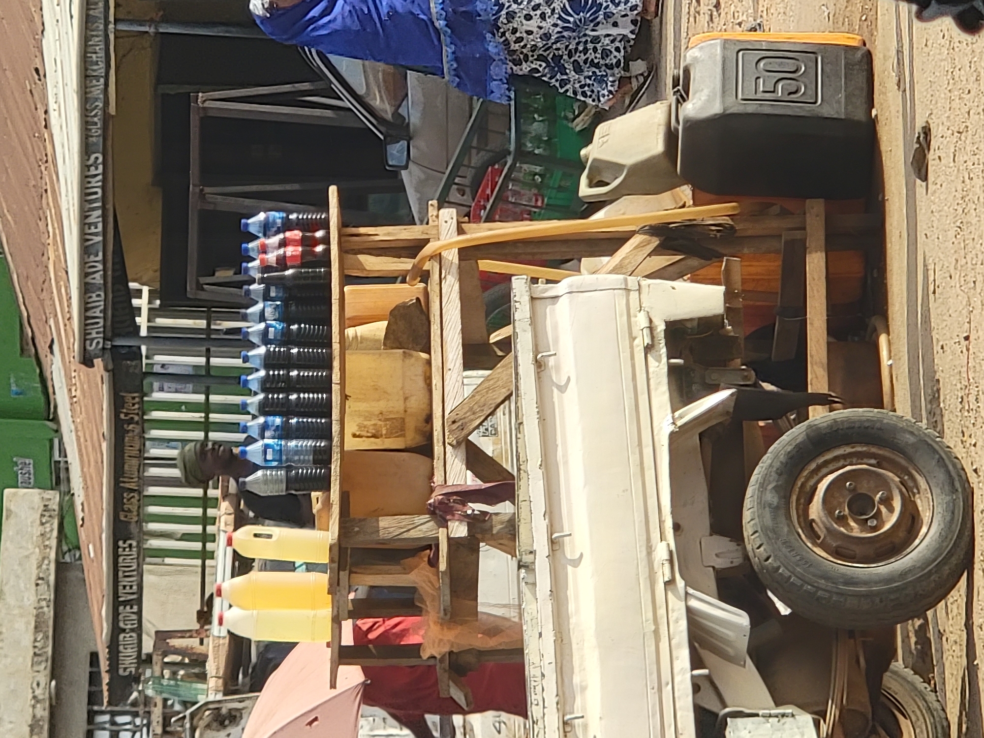 An outdoor shot depicts a street scene in Maiburiji, Nigeria. Stacked on a rickety wooden frame structure are water bottles, yellow plastic containers, and a black container labeled "50." The structure is mounted on a truck with a visible tire. A man stands nearby, and a building in the background displays signage for "SHUAIB ADE VENTURES, GLASS MERCHANTS, AL" and "SHUAIB-ADE VENTURES, Glass Aluminiums Steel." Clothing or fabric is draped over the top of the wooden structure.