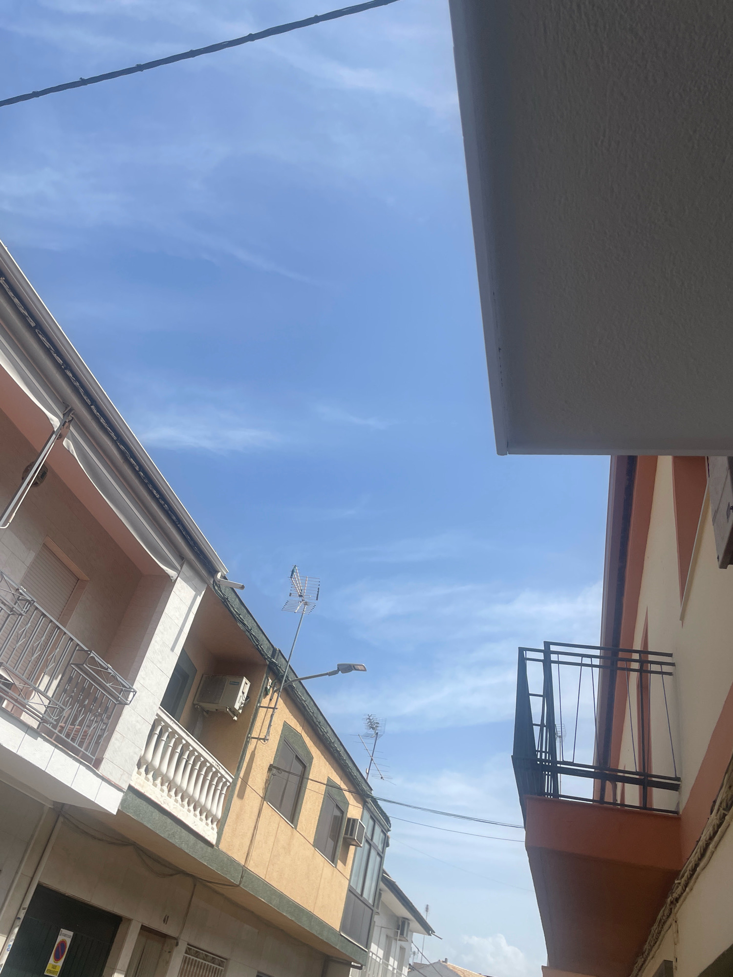The image shows an upward view of buildings against a bright blue sky with wispy clouds. On the left, a section of a building with a white and light pink facade is visible, featuring a balcony with a metal railing and a white awning. Further to the right, another building with a yellow facade is prominent, displaying windows, an air conditioning unit, and a balcony with a white balustrade. A street lamp and a television antenna are also attached to this building. On the far right, a building with an orange and white facade is partially visible, with a dark metal balcony. Electrical wires run across the sky, strung between the buildings. The scene is likely from a residential area in the city of Atarfe, Spain, given the architectural style and the presence of elements common to Mediterranean urban landscapes. No people or animals are visible in the image.