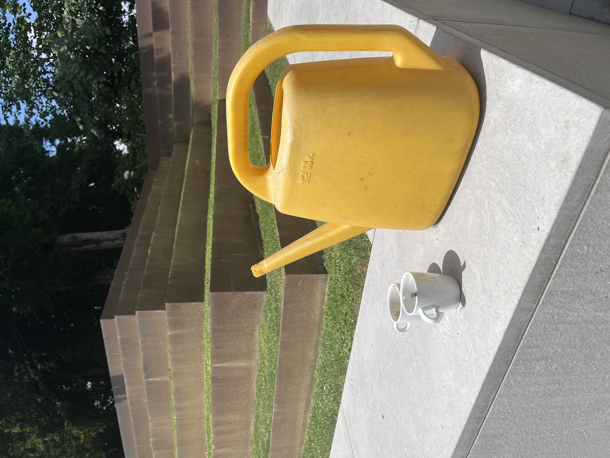 The image shows a bright yellow watering can with a capacity of 12 liters positioned on a concrete surface. Next to the watering can are two white mugs, one slightly in front of the other, casting distinct shadows on the concrete. The scene is set outdoors, with a background of tiered structures covered in grass and concrete. Trees and foliage are visible on the left side, with a blue sky peeking through. The lighting suggests it is daytime, likely sunny, as indicated by the strong shadows. The overall impression is one of a tranquil outdoor setting, possibly a garden or park area. There is no visible text on the mugs, but the watering can has "12L" embossed on its side. The image is oriented vertically.