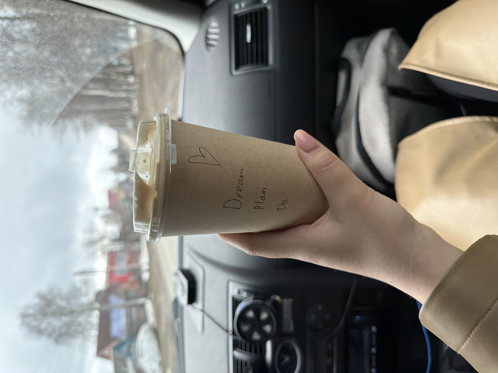 A hand holds a disposable coffee cup with the handwritten words "Dream. Plan. Do." and a heart symbol on it. The cup is in the interior of a car, as evidenced by the dashboard, vents, and other car features visible in the background. Outside the car window, a blurry view of a street scene can be seen. The location appears to be Votkinsk, Russia, based on the architectural style of the buildings and the wintry, overcast weather conditions. In the background, there are two light-colored handbags and what looks to be a gray-and-white sneaker, placed on the right passenger seat.