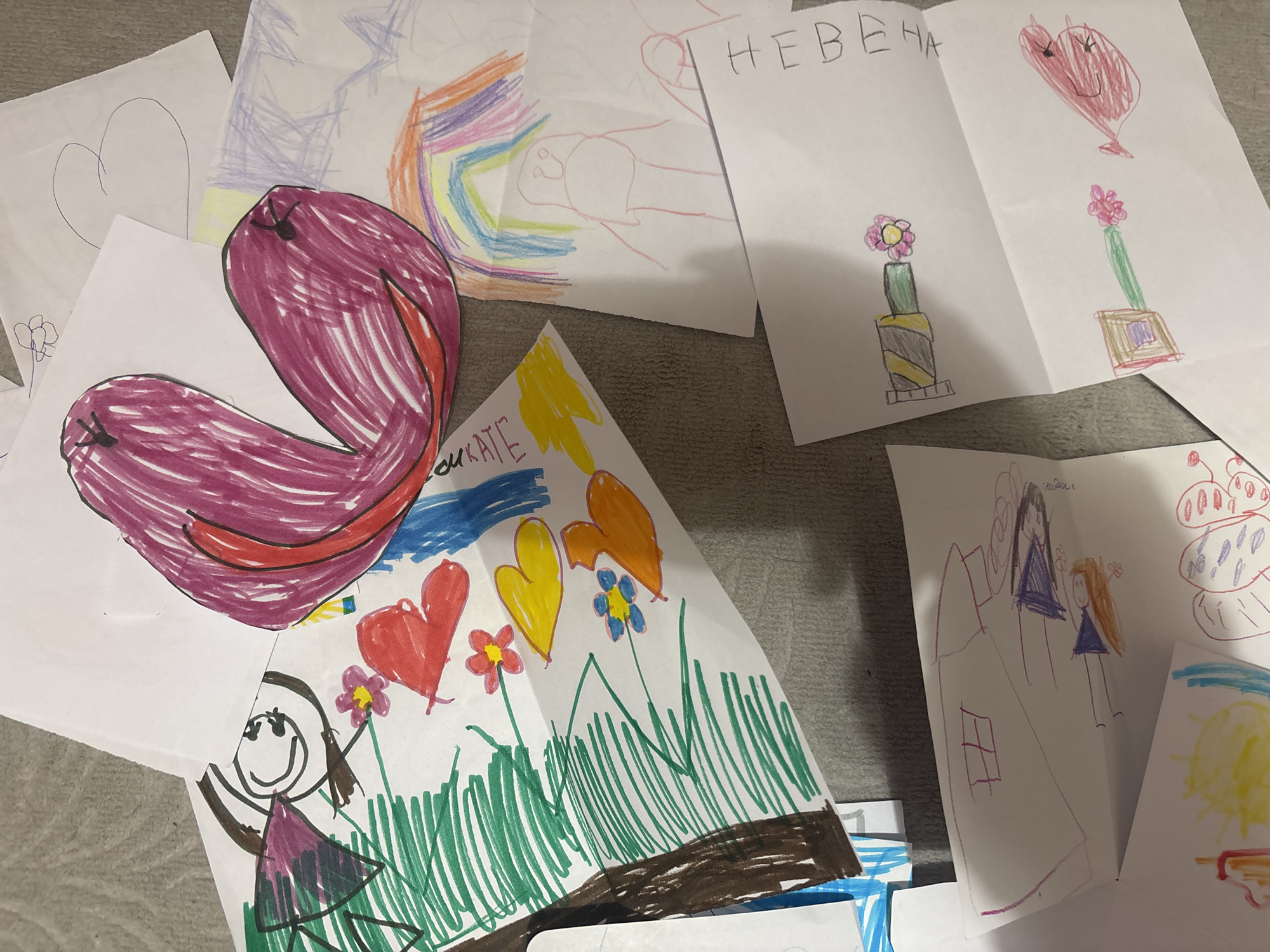 The image depicts a collection of children's drawings scattered on a surface. The drawings feature various subjects including hearts, flowers, people, a rainbow, and abstract shapes. One drawing includes the Cyrillic word "НЕВЕНА" (Nevena). The surface appears to be a floor or table. The location in Plovdiv, Bulgaria, cannot be determined from the visual information provided.