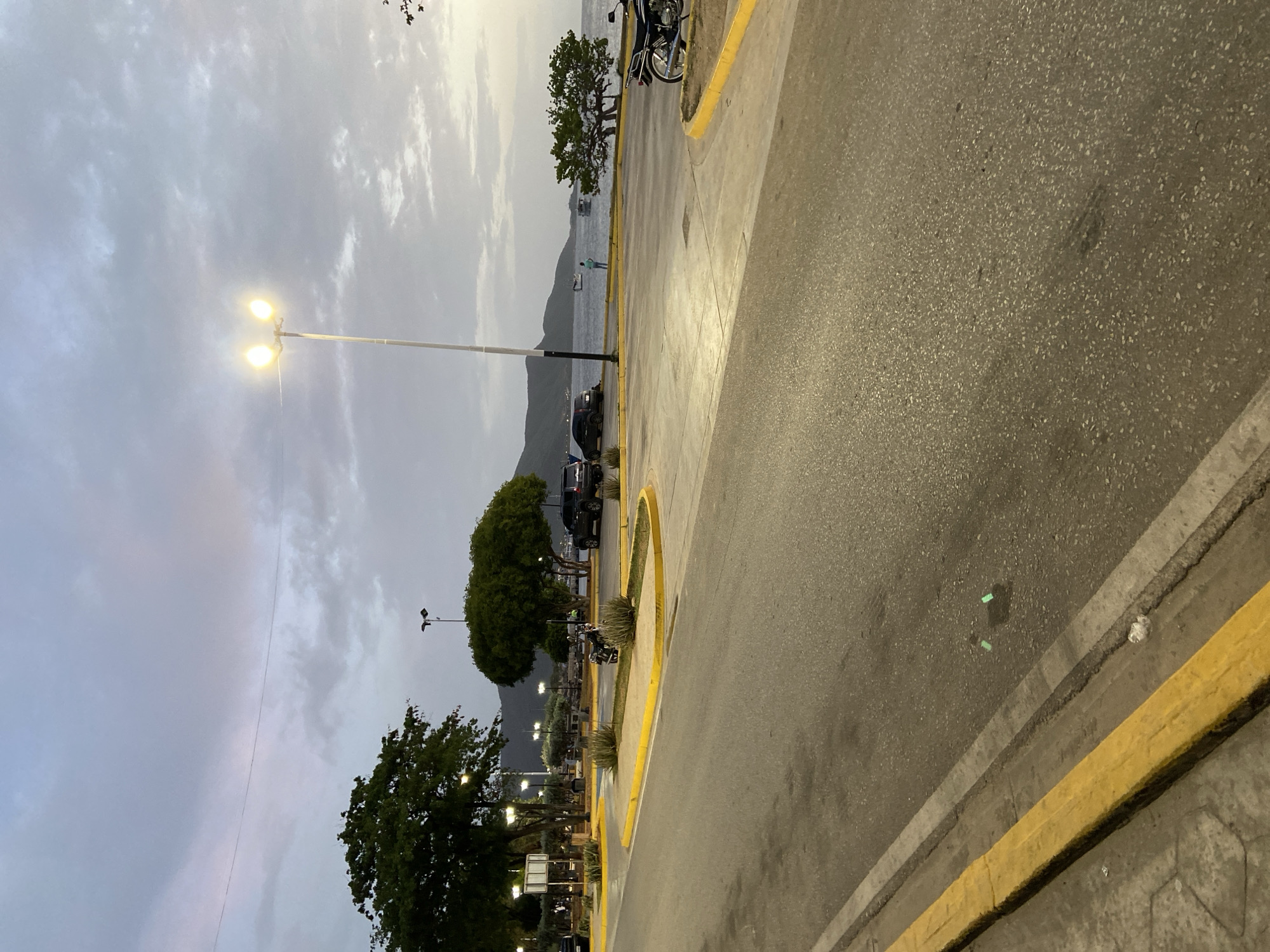 The image depicts a coastal road in Juan Griego, Venezuela, at dusk. A paved road with yellow and gray curbs curves through the scene. On the right side of the road, a parked motorcycle is visible. Across the road, a paved area with parked cars, including a black SUV, is present. Beyond the paved area, a grassy area with trees and streetlights is situated. In the background, the ocean stretches out, with several boats and a distant shoreline featuring hills. The sky is overcast with hues of gray and purple, characteristic of twilight. A lone figure stands near the water's edge, observing the sea. The overall atmosphere is calm and serene.