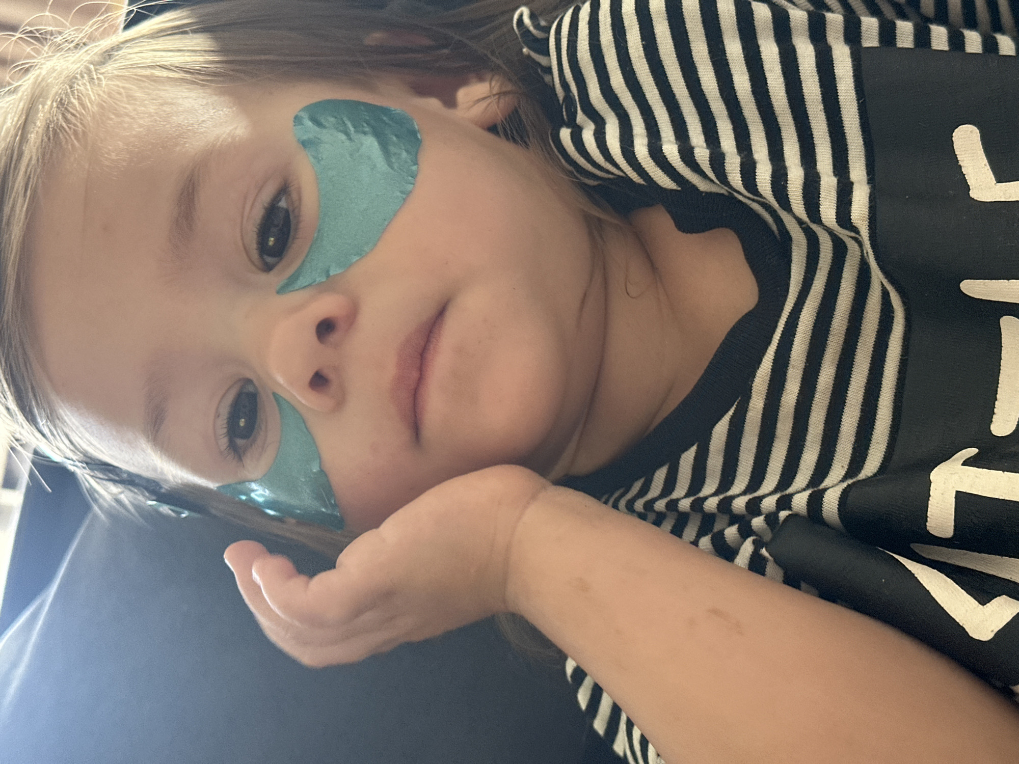 A close-up, waist-up, indoor, eye-level shot shows a young child with light brown hair and fair skin. The child is wearing a black and white striped t-shirt. Two teal-colored under-eye masks are applied to the skin below the child's eyes. The child's left hand is placed on their cheek, obscuring part of their face. The background is dark and out of focus, with a slight hint of vertical lines suggesting a surface. The image provides no information about the city of Kharnavy, Russia, or any specific environmental details.