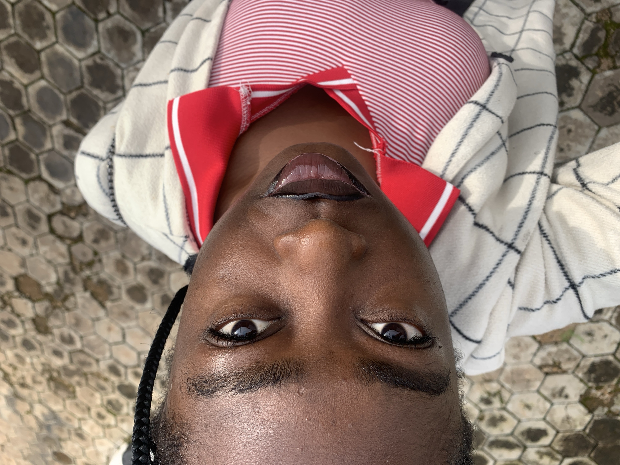 A close-up, high-angle, vertically oriented shot shows a young Black person from the shoulders up, looking upwards towards the camera. The person's skin is dark brown, with visible pores and some sheen from natural oils or product. Their eyebrows are dark and neatly shaped. Their eyes are dark brown, with long lashes and a slight sparkle. The left eye is more visible and the iris is clearly discernible. The lips are full, with a glossy, reddish-brown lipstick that creates a wet appearance. A single dark braid runs from the left temple area and disappears out of frame to the left. The person is wearing a red and white striped collared shirt, and over that, a cream-colored cardigan with a black plaid pattern. The background is an outdoor, paved surface with hexagonal-shaped stones, some of which appear weathered or slightly mossy, suggesting an urban or semi-urban environment. The lighting suggests it is daytime. There is no specific information that allows for determination of location within Gura Top, Nigeria.