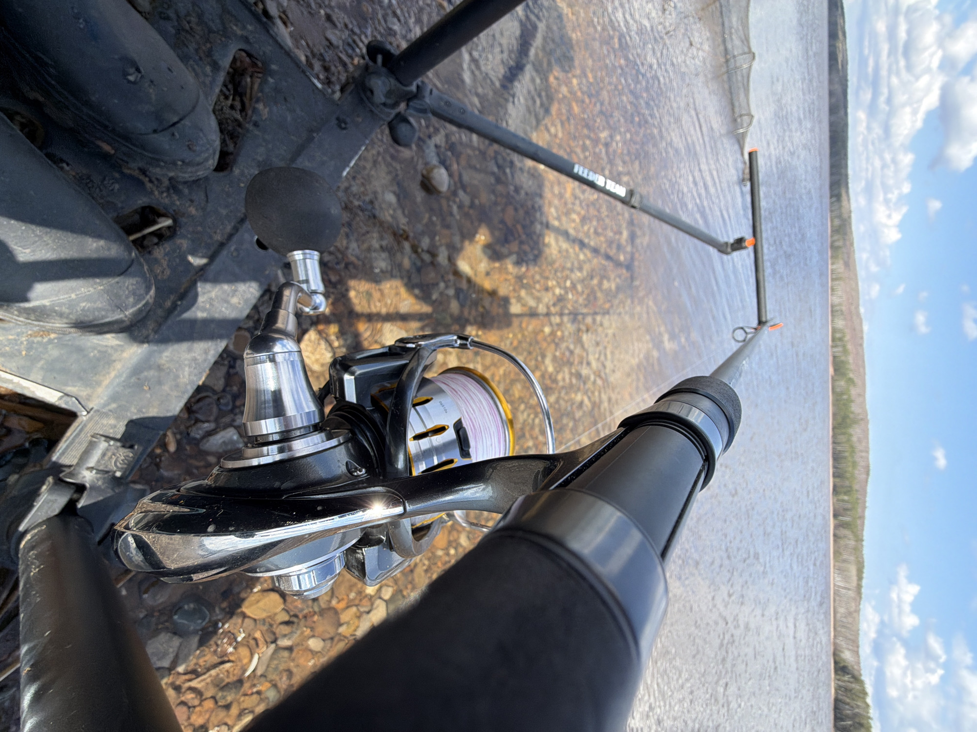 The image captures a first-person perspective of a fishing setup on a rocky or gravelly bank beside a wide body of water, likely a river or lake, in Chaikovsky, Russia. In the foreground, a pair of black boots is visible on the ground.
The primary subject is a fishing rod and reel, held in a rod stand. The fishing reel features a silver and black design with a visible spool wound with light pink or white fishing line. The rod itself extends towards the water. The rod stand, black with orange accents, is partially submerged in the shallow water near the bank, with the text "FEEDER TEAM" clearly visible on one of its legs, indicating the type of fishing being done.
Further out in the water, a dark-colored keepnet is partially submerged, suggesting fish may be retained or are intended to be. The water surface reflects the bright sunlight and appears calm, with small ripples. The opposite bank or distant shore is visible in the background, appearing as a long, dark line of land, possibly forested. Above, the sky is clear blue with scattered white clouds, indicating a sunny day, likely morning or afternoon. The overall scene suggests a calm and focused fishing activity.