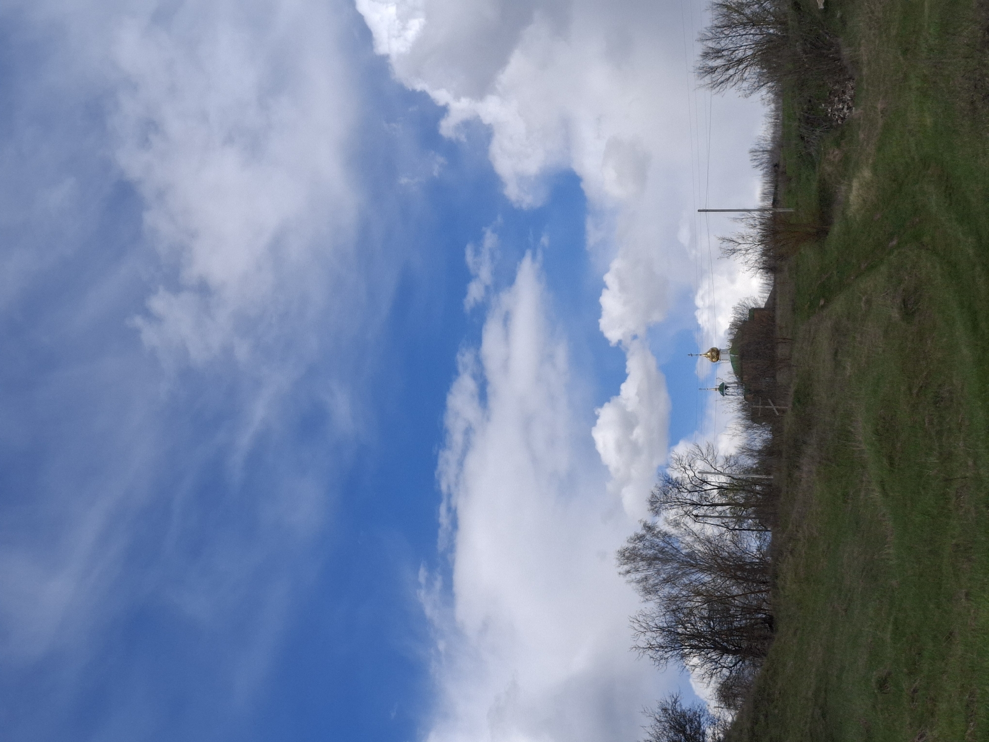 The image, when viewed in its correct orientation, presents a serene rural landscape under a vast, partly cloudy sky. The upper portion of the frame is filled with a bright blue sky, adorned with scattered white and grey clouds, ranging from fluffy cumulus formations to more diffuse, wispy streaks, indicating daytime.
The lower half depicts a gently undulating grassy hillside or embankment, covered in vivid green grass, suggesting either spring or early summer. Along the slope and crest of the hill are numerous deciduous trees and shrubs, currently bare with visible branches, characteristic of early spring before the foliage emerges.
On the horizon line, notable features include the golden, onion-shaped dome of a church or chapel, topped with a cross, partially obscured by the landscape. A wooden utility pole with power lines running horizontally across the mid-ground is also visible. A subtle path or track can be seen traversing the grassy slope on the right side. A small pile of rocks or debris is noticeable amongst the sparse vegetation.
The scene captures a quiet, open expanse, free of human activity, likely in the Елец-Маланино region of Russia, marked by its natural beauty and the presence of an Orthodox religious structure. No text is visible in the image.