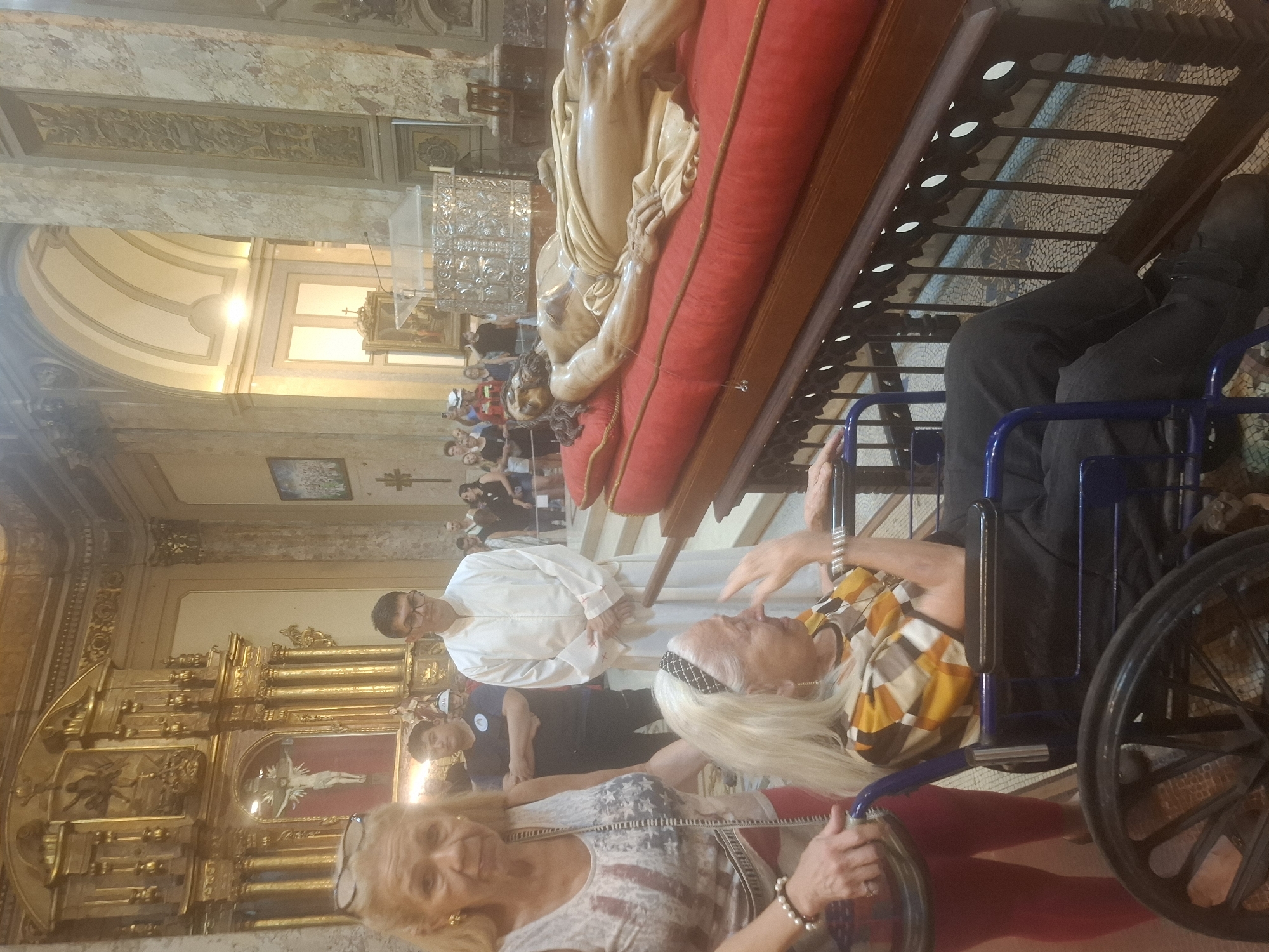 The image shows the interior of a church in Buenos Aires, Argentina, possibly during a religious procession or event. A group of people are visible, with a focus on two elderly women in the foreground. One woman is wearing a shirt with an American flag design and a pair of glasses is hanging off of her chest; the other is partially visible in a wheelchair. A figure of Jesus Christ is on a red platform, lifted above the crowd, and a priest in a white robe stands nearby. The interior of the church is ornate with golden accents and architectural details.