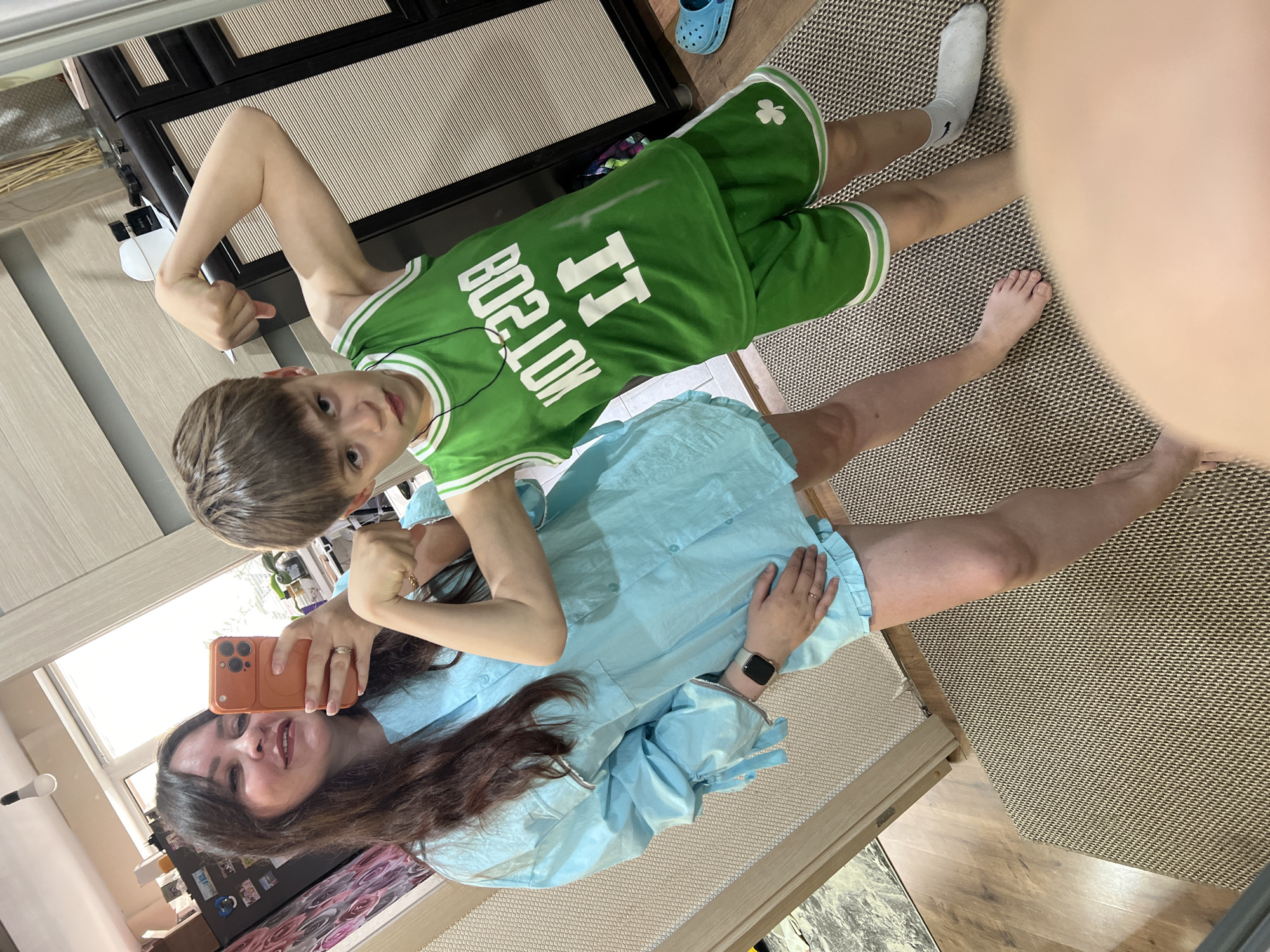 A woman and a boy are in a residence. The woman is taking a selfie with an orange smartphone. The boy is wearing a green Boston Celtics jersey and shorts. He flexes his bicep. The floor is covered with a brown and beige carpet. A white sock is on the boy's left foot and his right foot is bare. A blue Croc is in the background. A dark-colored TV stand is against the wall. The boy has ear buds in. The woman is wearing a light blue garment and a watch. Magnets are on a refrigerator. The city is not discernible in the image.
