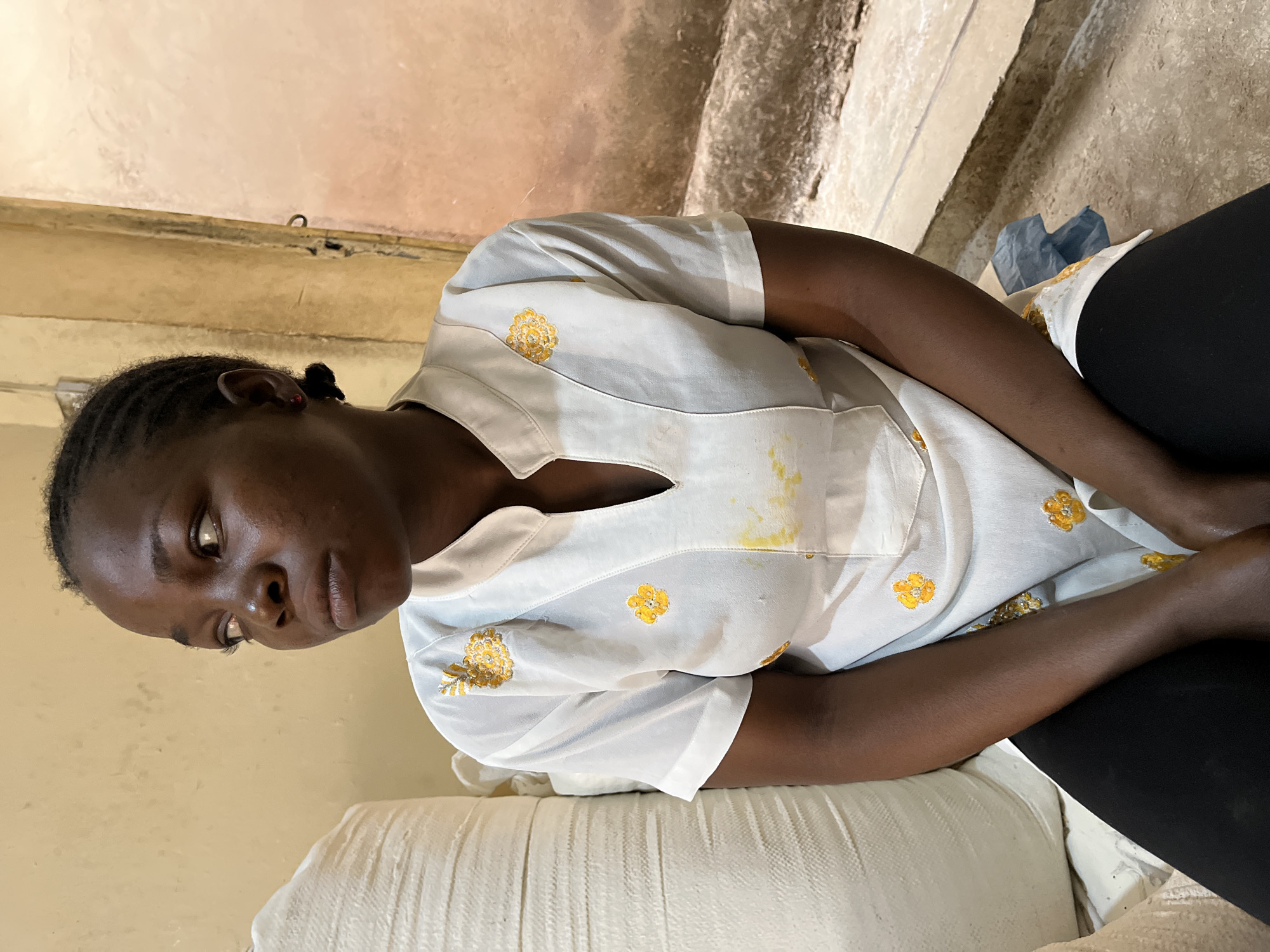 A young Black woman sits indoors, appearing pensive or sad. She is wearing a light-colored shirt with yellow floral designs, and black leggings. She is sitting on a white cushion or pillow. The location appears to be a simple, possibly unfinished interior space. The photograph was taken in Jos, Nigeria.