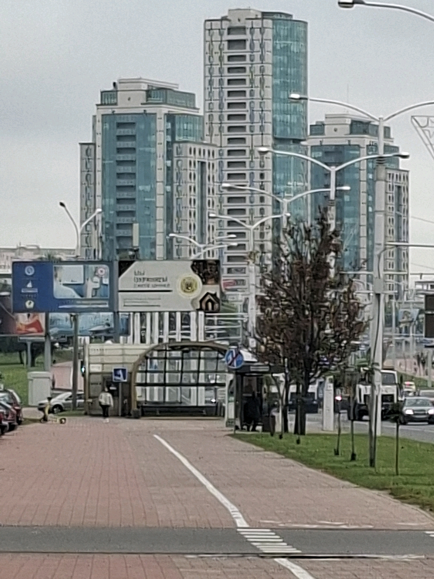 The image depicts a street scene in Minsk, Belarus, characterized by modern high-rise buildings and urban infrastructure. In the foreground, a red brick pedestrian walkway with a white dashed line marking a crosswalk leads towards a bus stop shelter. A woman in a light-colored hooded jacket and dark pants is walking on the walkway, approaching an entrance with a blue handicap symbol. To the left of the walkway, several cars are parked along the side of the road, and a yellow scooter is also visible.
In the midground, a bus stop shelter with glass panels is situated next to the walkway, with people visible inside. Adjacent to the bus stop, a cluster of billboards displays various advertisements. Behind these, a modern building complex dominates the skyline. These buildings are characterized by their light-colored facades and extensive use of blue glass, featuring multiple floors with repetitive window patterns. Some of these buildings have a stepped or tiered design, with sections set back from the main structure.
The background reveals more of the cityscape, with additional tall buildings and a street with vehicles, including trucks and cars, driving on it. Streetlights with distinctive curved arms are visible throughout the scene, interspersed with trees, some of which appear to be bare of leaves, suggesting it might be autumn or winter. The sky is overcast with a light gray color, contributing to a diffused lighting condition. The overall impression is one of a contemporary urban environment.