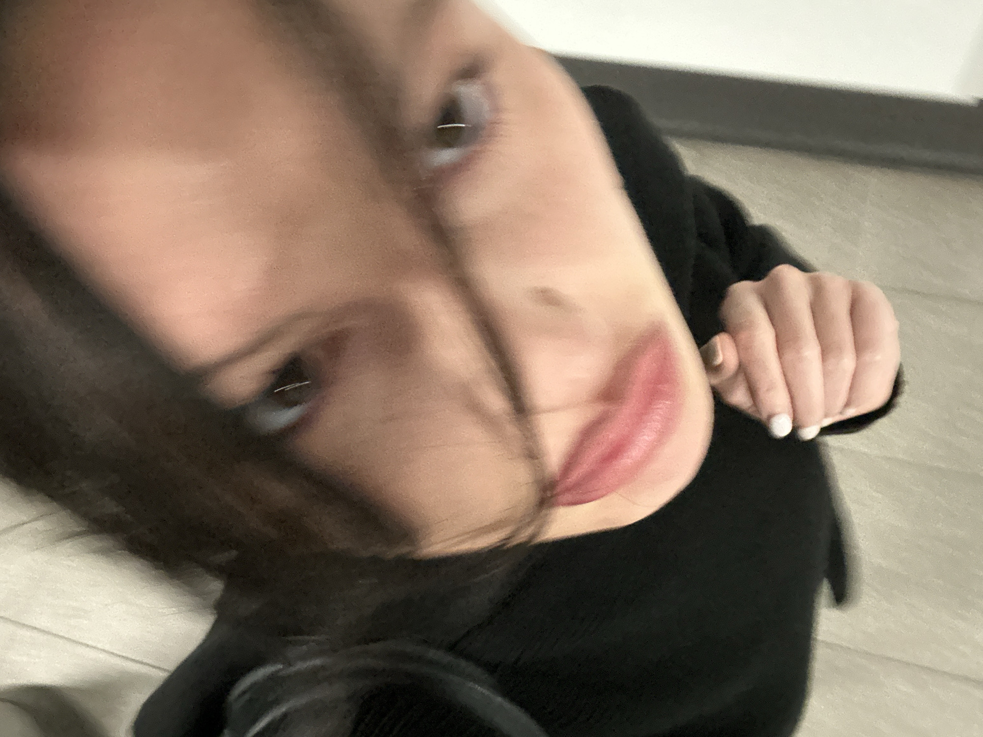 A close-up, slightly tilted selfie of a woman. The background is out of focus and appears to be a neutral-colored wall or floor. The woman has dark hair with some strands falling across her face, light skin, and pink lips. Her hand is visible on the right side of the frame with white nail polish. She is wearing a black top. The location is indeterminate as no landmarks are visible.