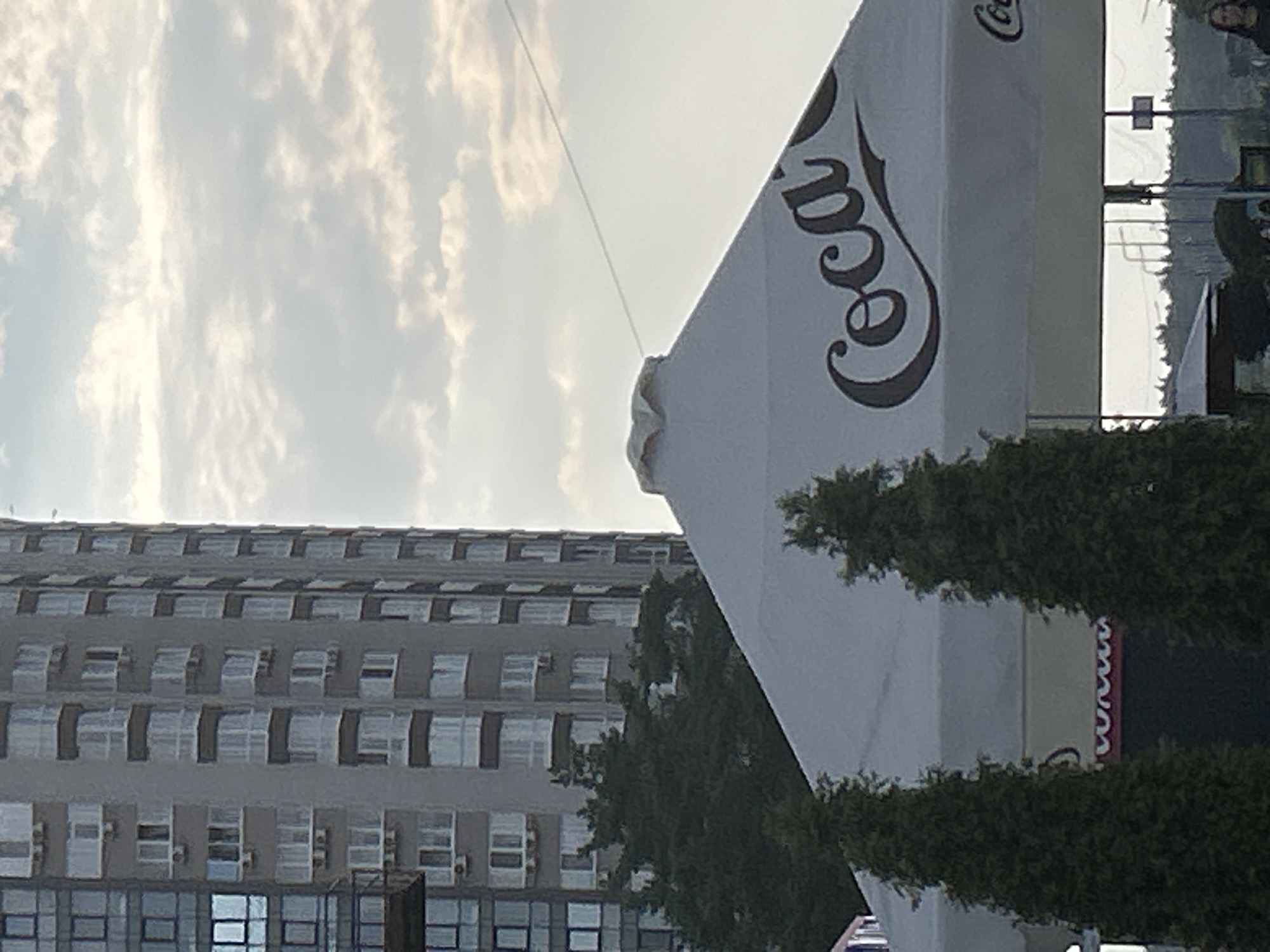 The image shows a tall, modern apartment building in Brovary, Ukraine, with a distinctive glass facade on the left side. In the foreground, a large white umbrella with the Coca-Cola logo is prominently displayed. Two evergreen trees stand in front of the umbrella. The sky above is cloudy, with a soft, diffused light. In the background, a line of trees and some structures are visible, suggesting a partially developed or suburban area. The overall scene captures an outdoor urban environment under a cloudy sky.