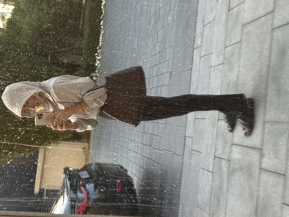 The image captures the reflection of an individual in a glass surface speckled with numerous small droplets, under strong, direct sunlight. The individual is presented in profile, facing left, wearing a light grey cropped hooded sweatshirt with the hood up, dark flared pants, and dark open-toed two-strap sandals. They hold a light-colored smartphone with both hands at chest height, angled as if taking a self-portrait. A dark, textured handbag is positioned at their hip. Multiple thin bracelets are visible on the individual's right wrist. The background features the rear of a dark-colored vehicle, likely an SUV or sedan, parked on a paved area to the left. To the right, green foliage is visible. The ground consists of a sidewalk paved with grey rectangular blocks. This environment is consistent with a residential or commercial street in Dubai, United Arab Emirates.